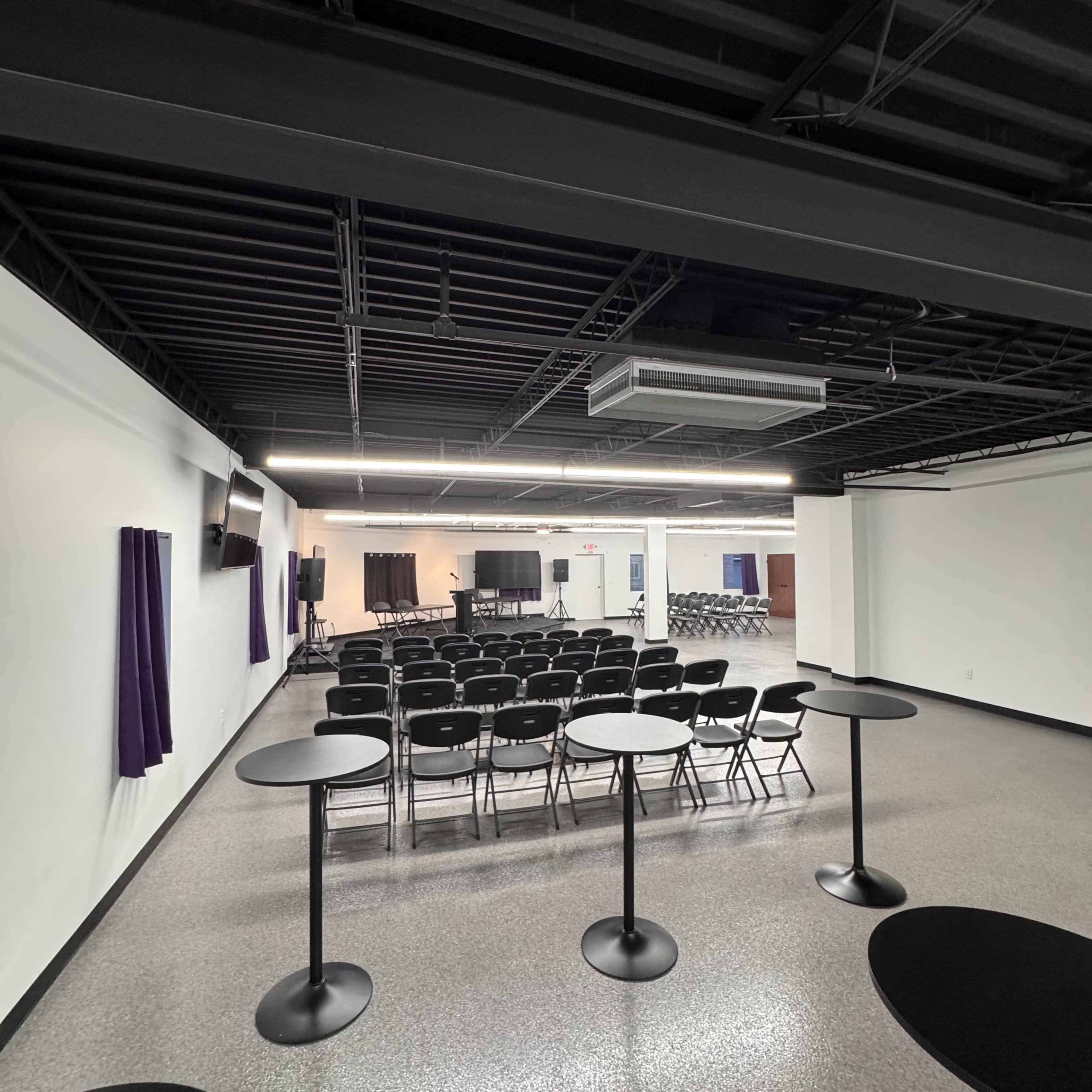 The image shows a spacious, well-lit event room with rows of chairs facing a presentation area, surrounded by tables.