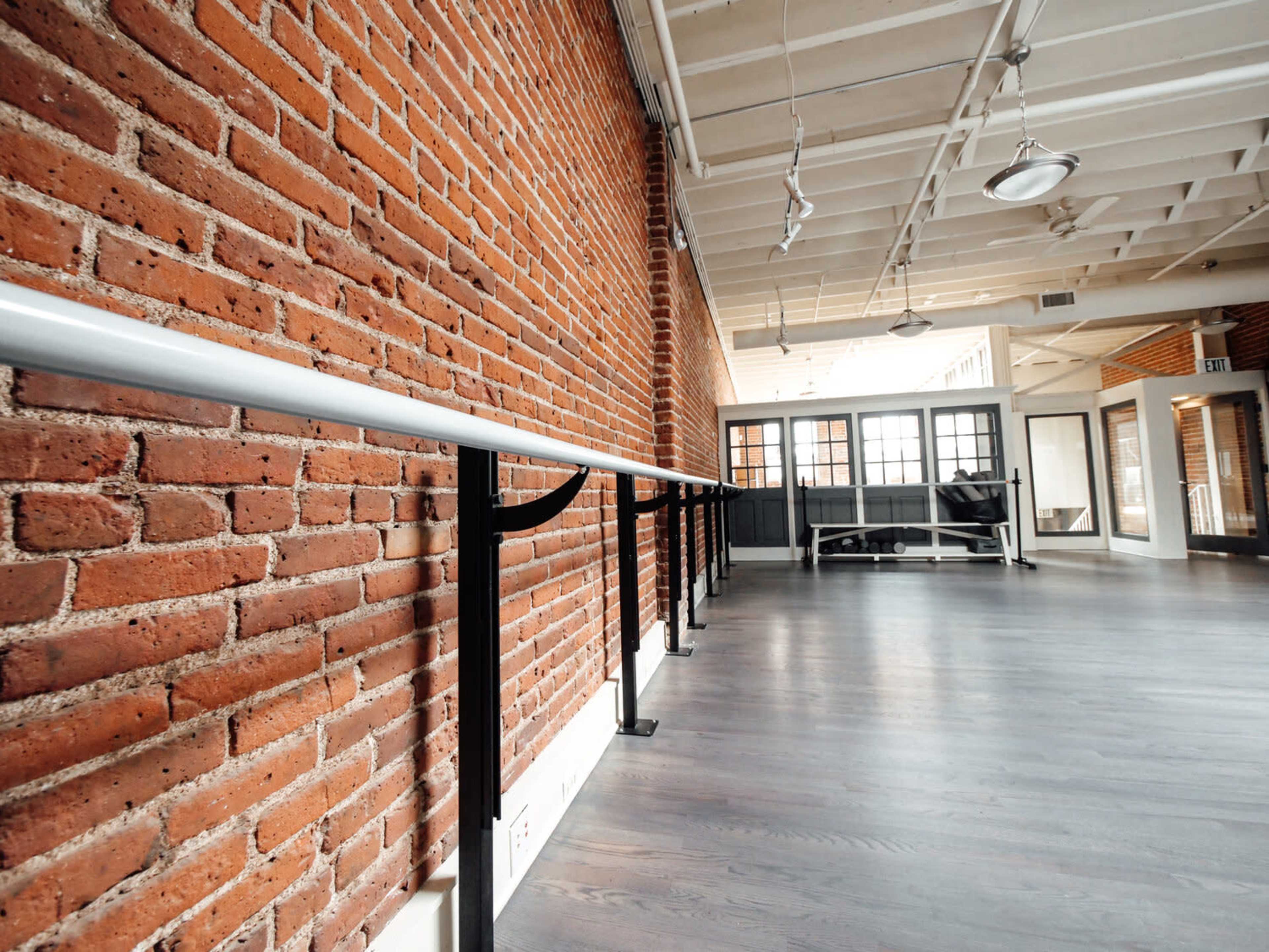 The image shows a spacious interior with a wooden floor, a brick wall, and a ballet barre mounted along one side.