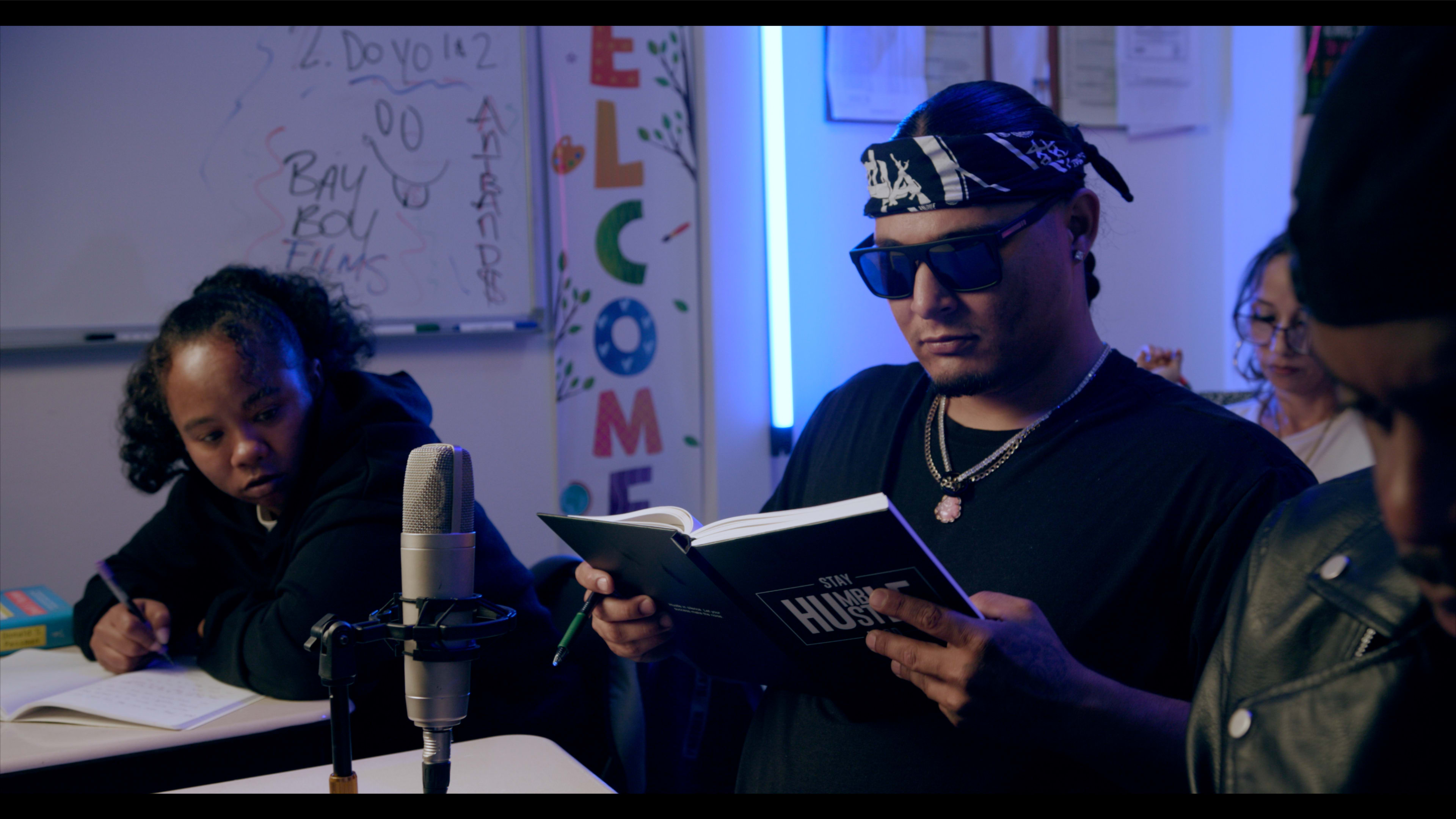A man wearing sunglasses and a bandana reads a book while a woman takes notes at a table in a room decorated with colorful signage.