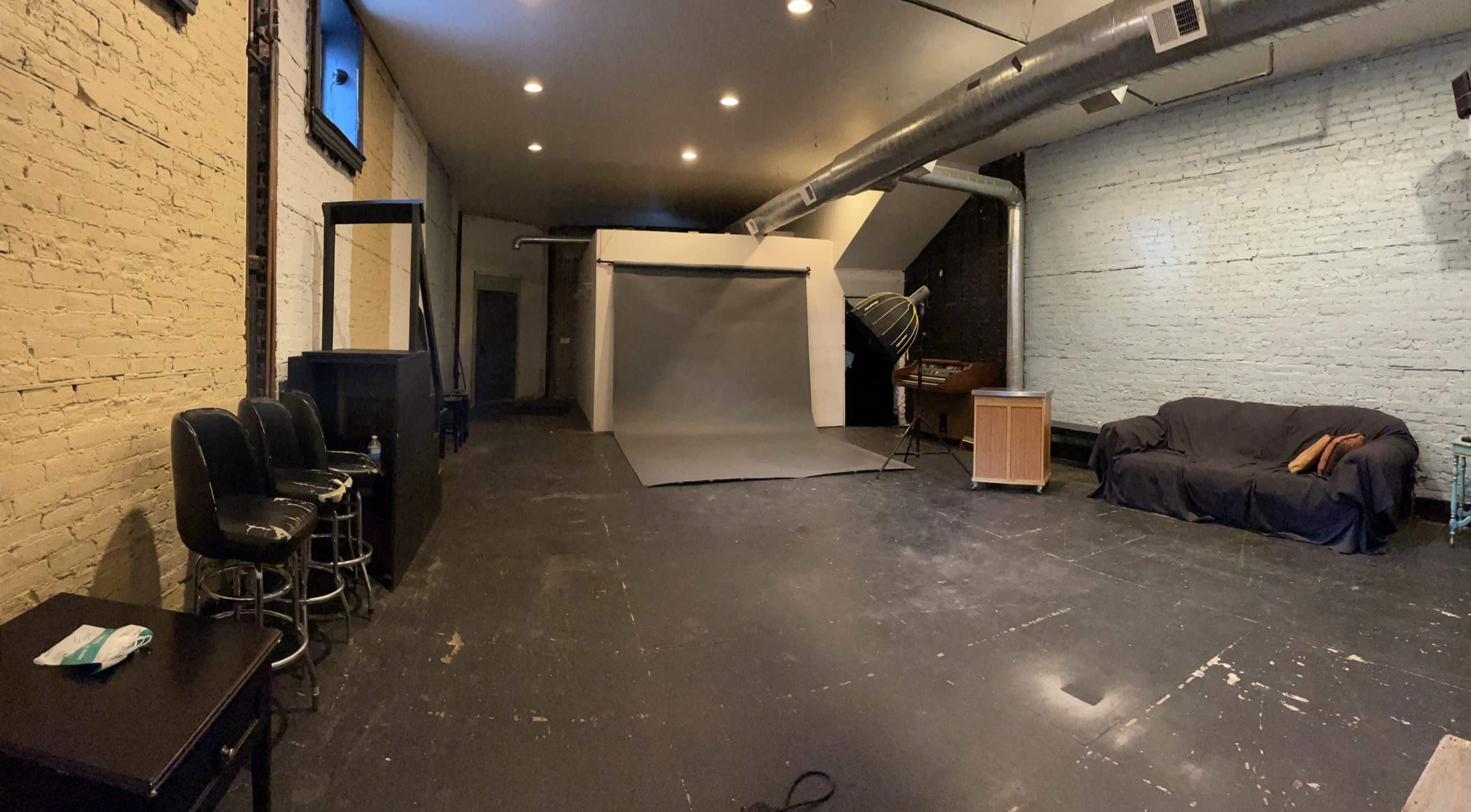 The image shows a spacious studio with bare walls, a gray backdrop set up for photography, a couch, a table, and several bar stools.