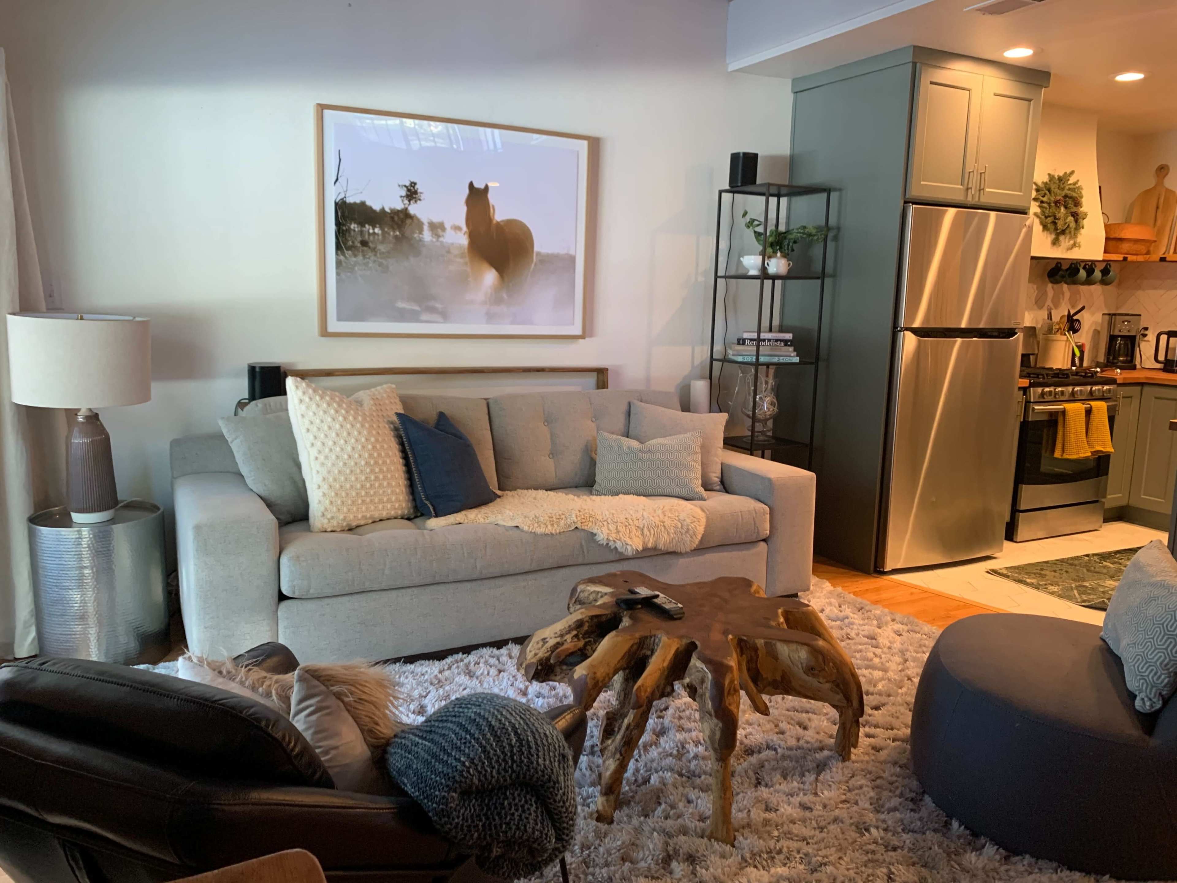 A cozy living room with a gray sofa, a wooden coffee table, and a kitchen area in the background, featuring stainless steel appliances.