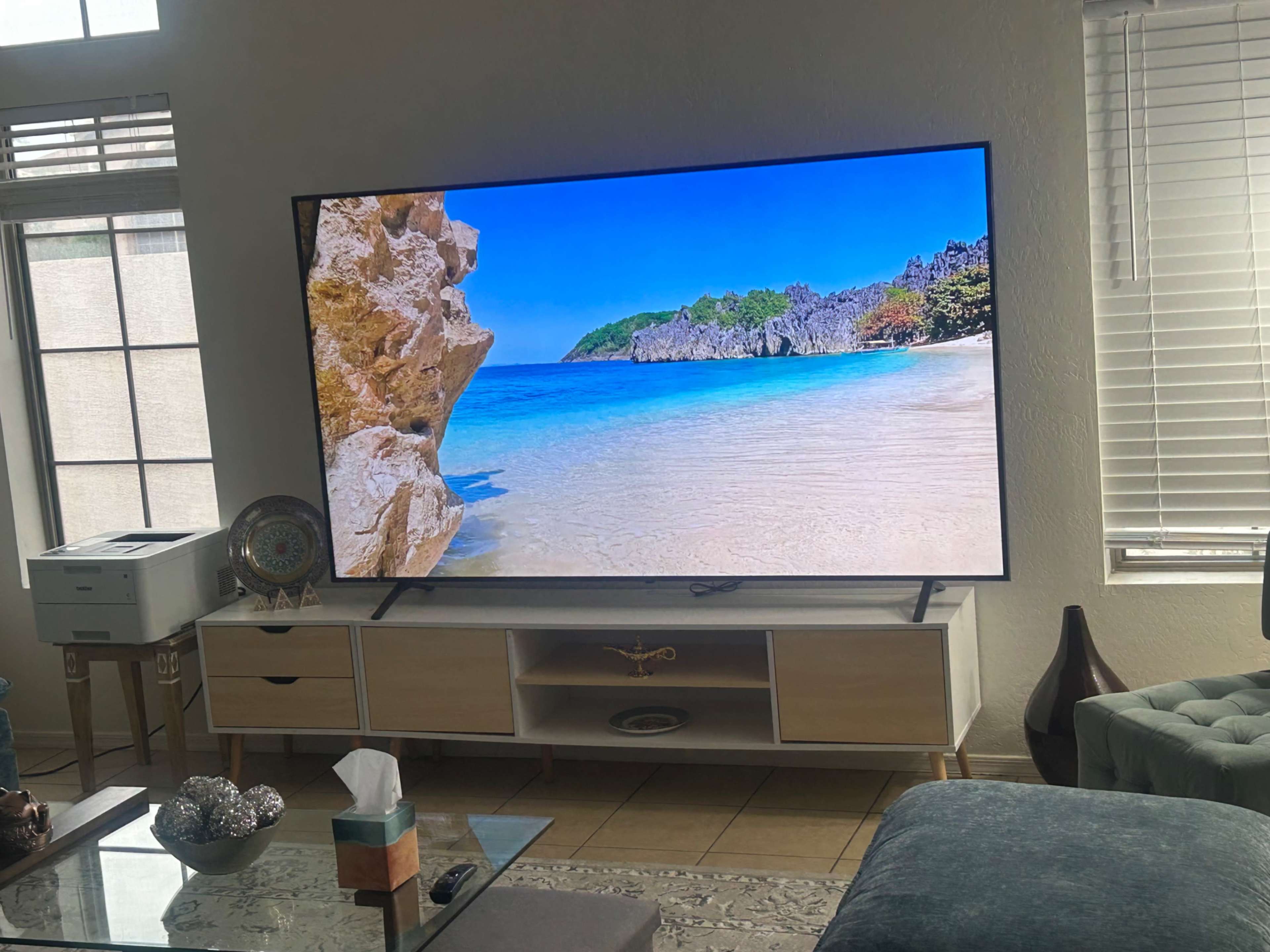 A large television displays a vibrant beach scene with clear blue water and rocky cliffs, positioned in a living room next to a printer and a coffee table.