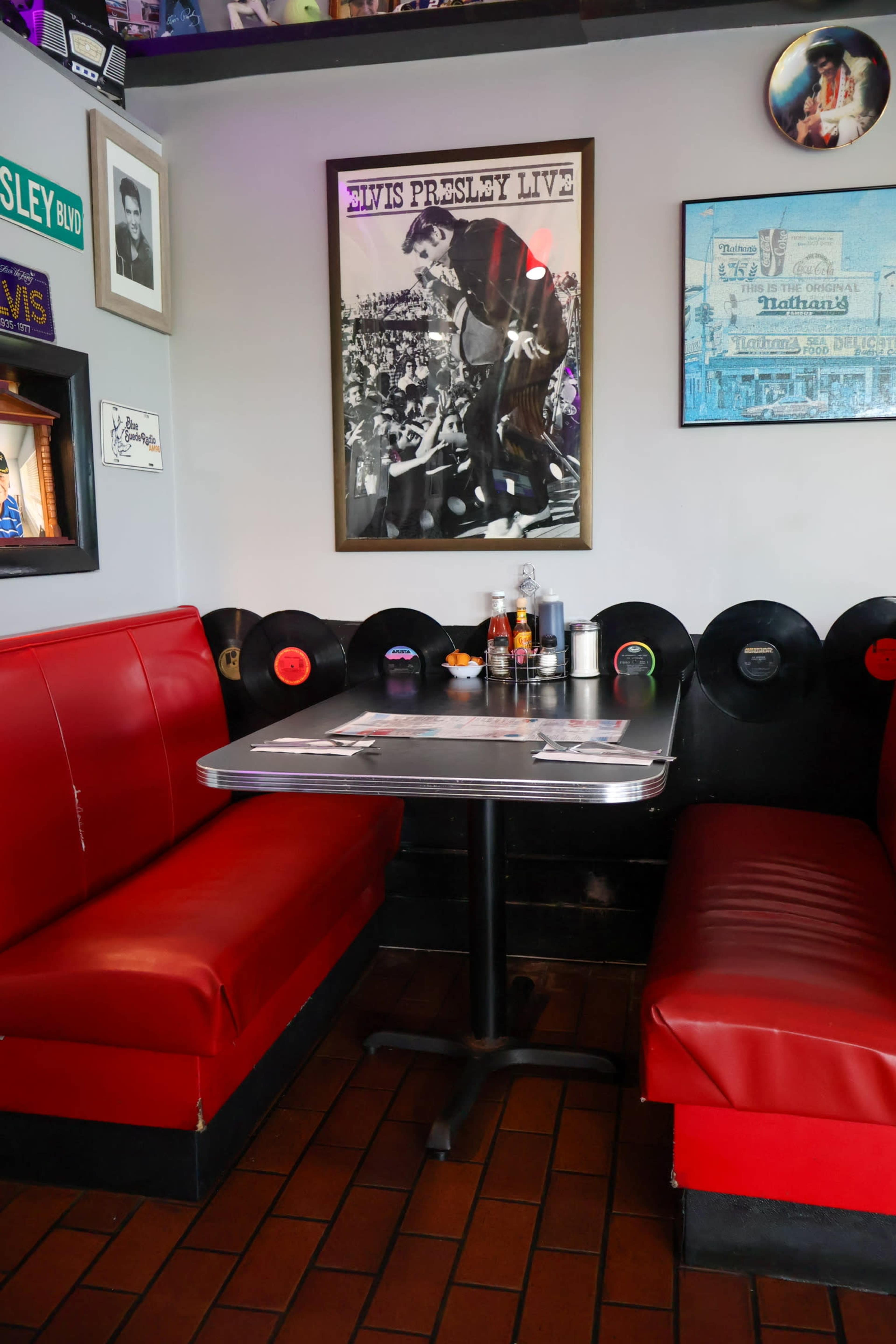 A red booth with a black table is situated in a diner adorned with Elvis Presley memorabilia and vintage records on the walls.