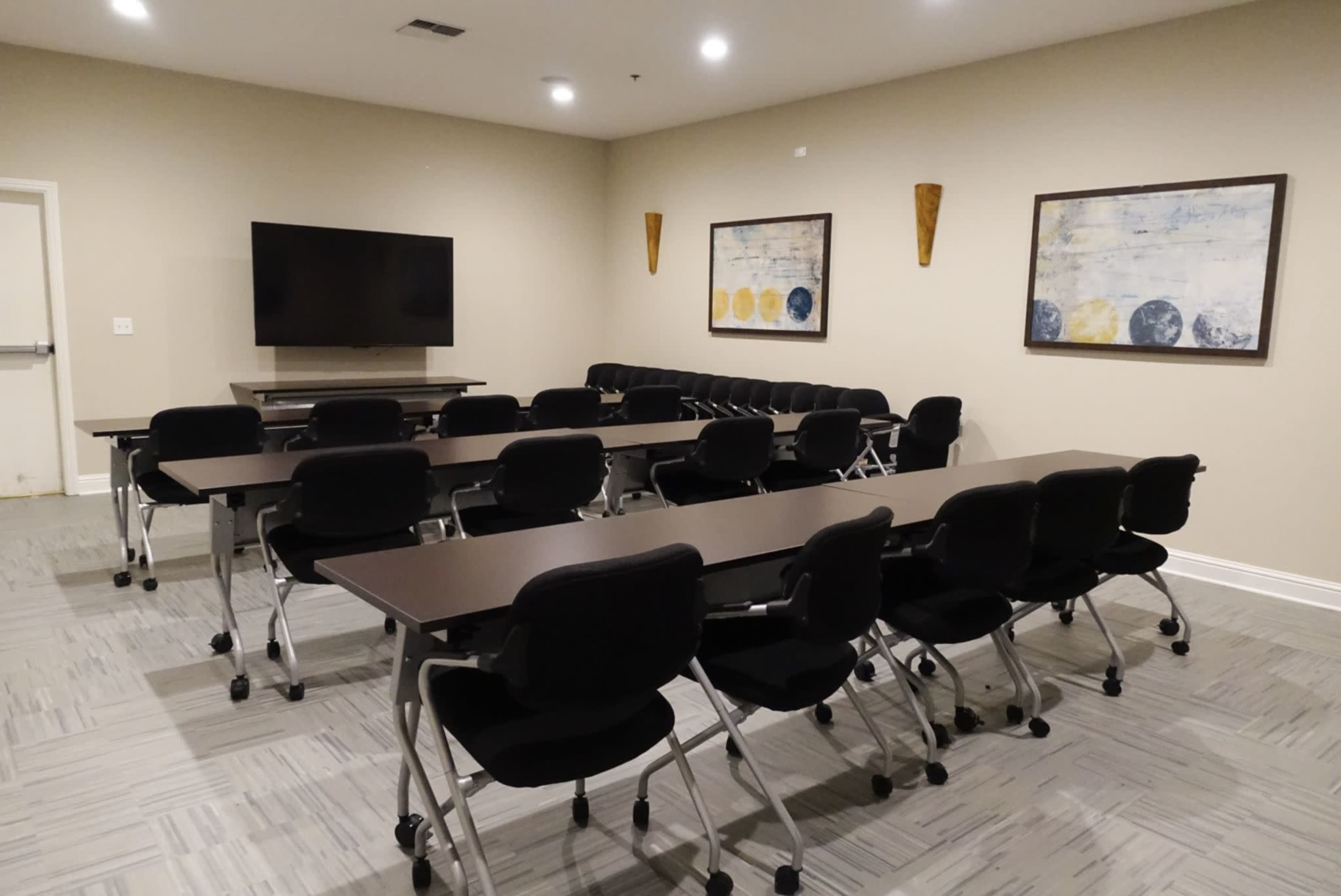 A conference room equipped with multiple tables and chairs arranged for a meeting, featuring a television on the wall and two framed artworks.