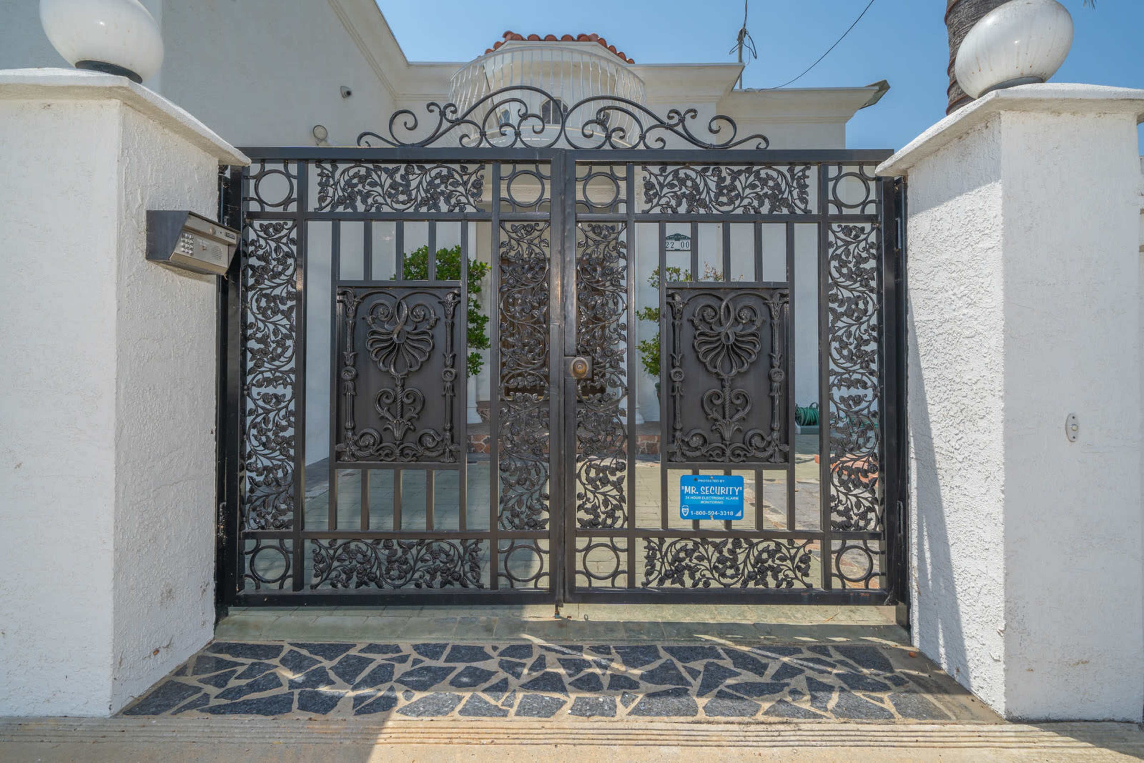 An ornate black metal gate with decorative designs, leading to a property with a stone pathway and greenery visible beyond.