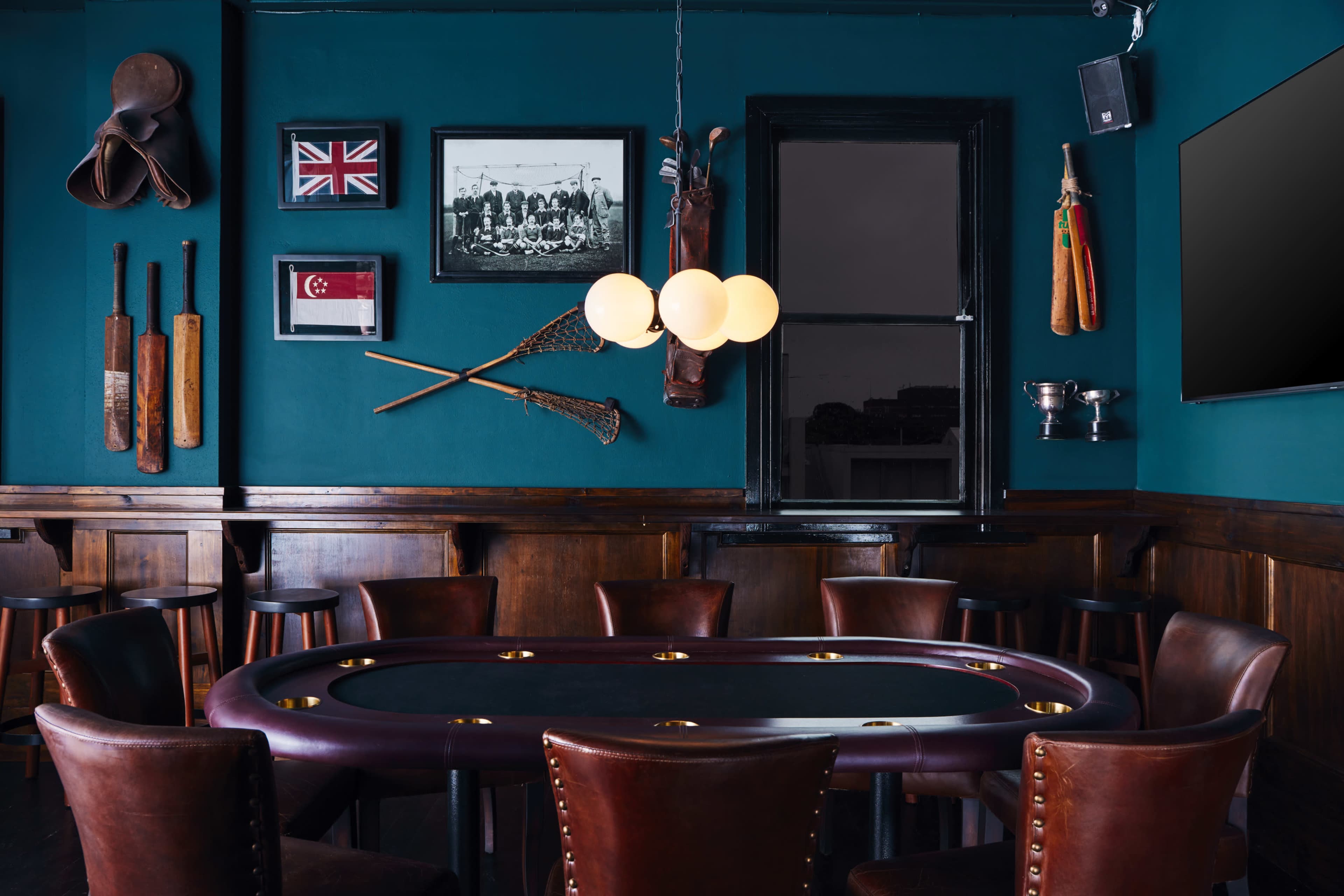A dimly lit room featuring a round poker table surrounded by leather chairs, with framed sports memorabilia and a large screen on the walls.