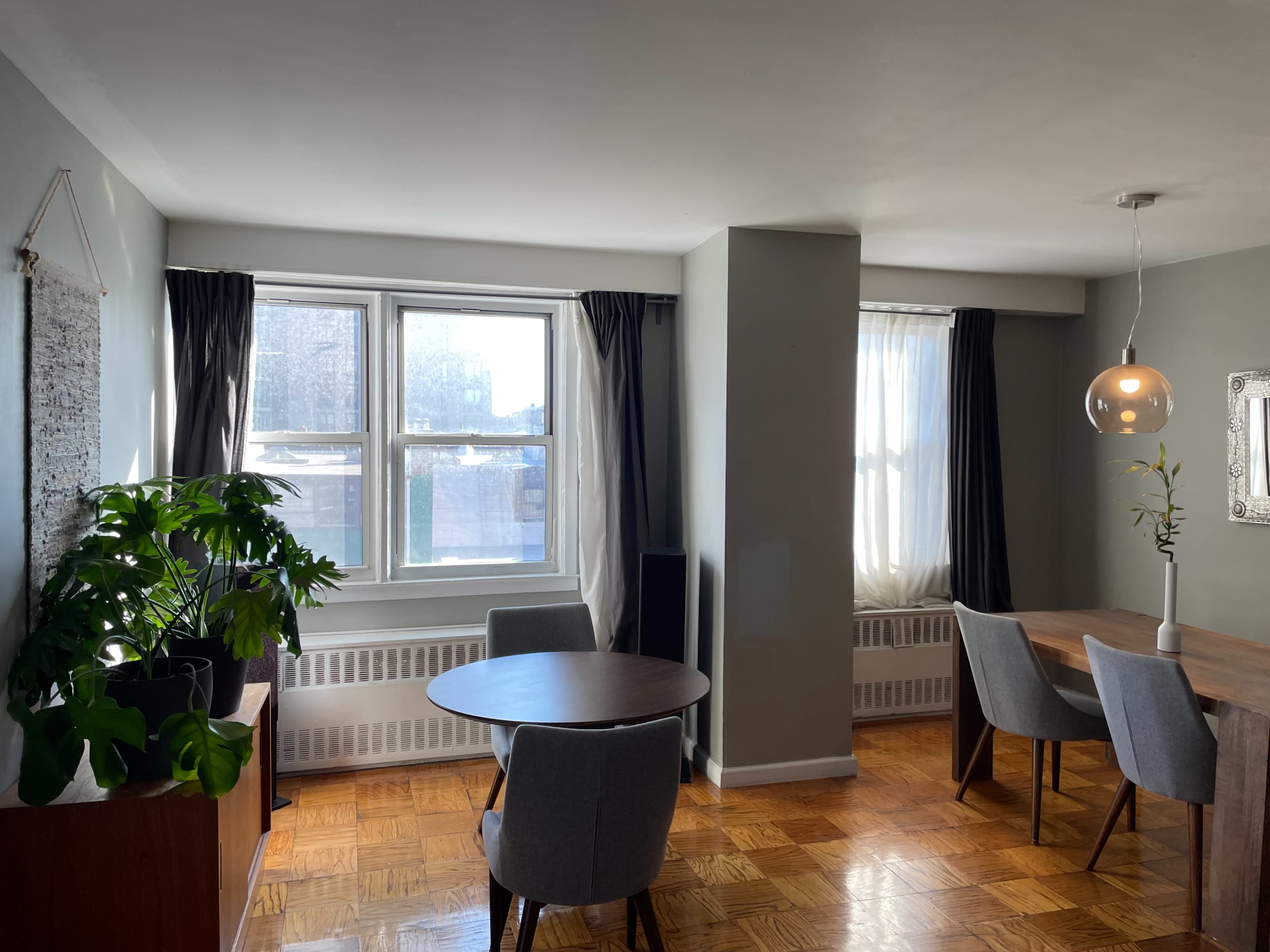 A modern dining area features a round table with gray chairs, a wooden dining table, and large windows with gray curtains.