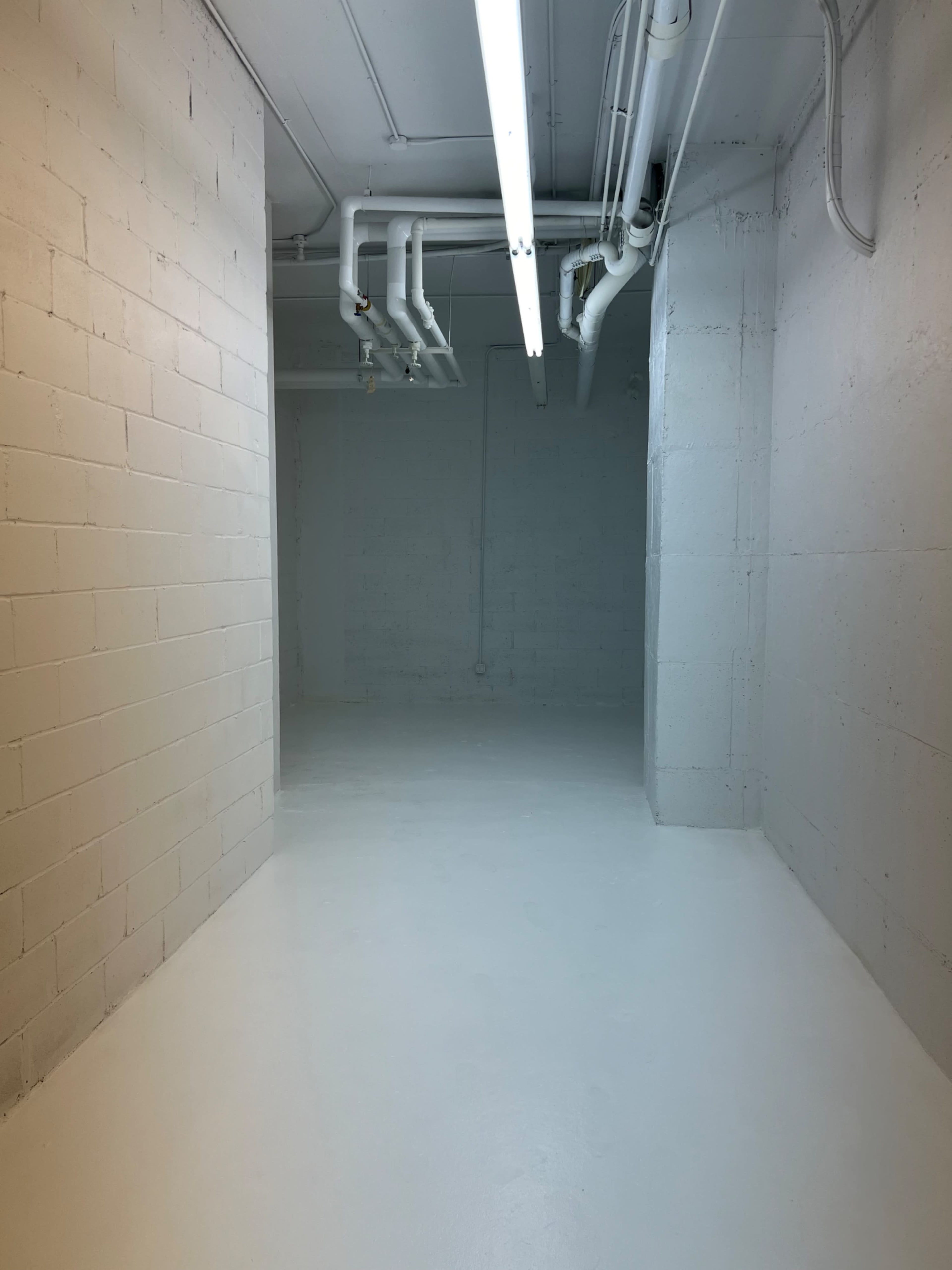 A large, empty room with white walls and a concrete floor, illuminated by overhead fluorescent lighting and featuring exposed pipes along the ceiling.