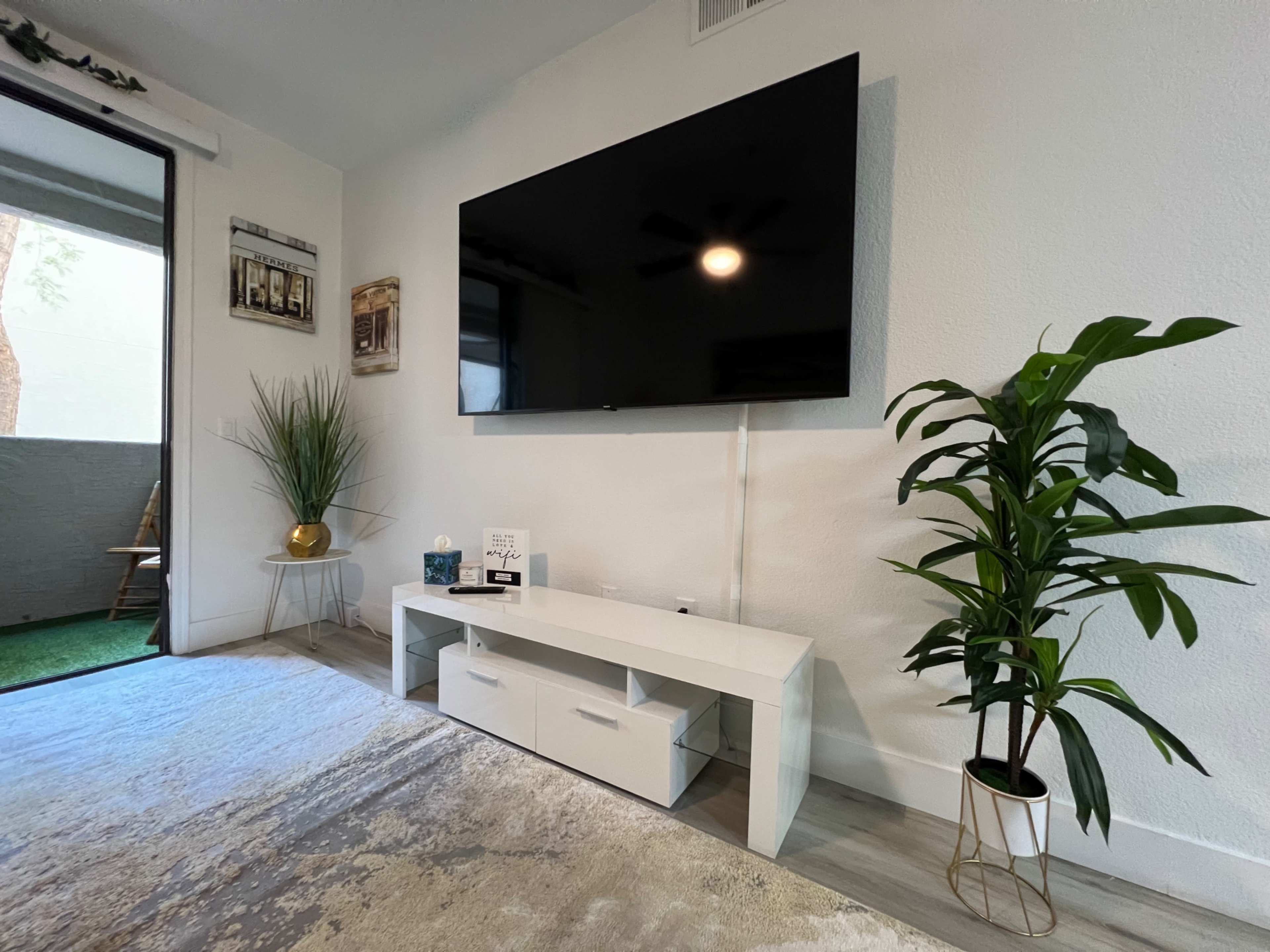 A modern living room features a large flat-screen TV mounted on a white wall above a sleek white TV stand, accompanied by a potted plant and a glass door leading to a small outdoor area.