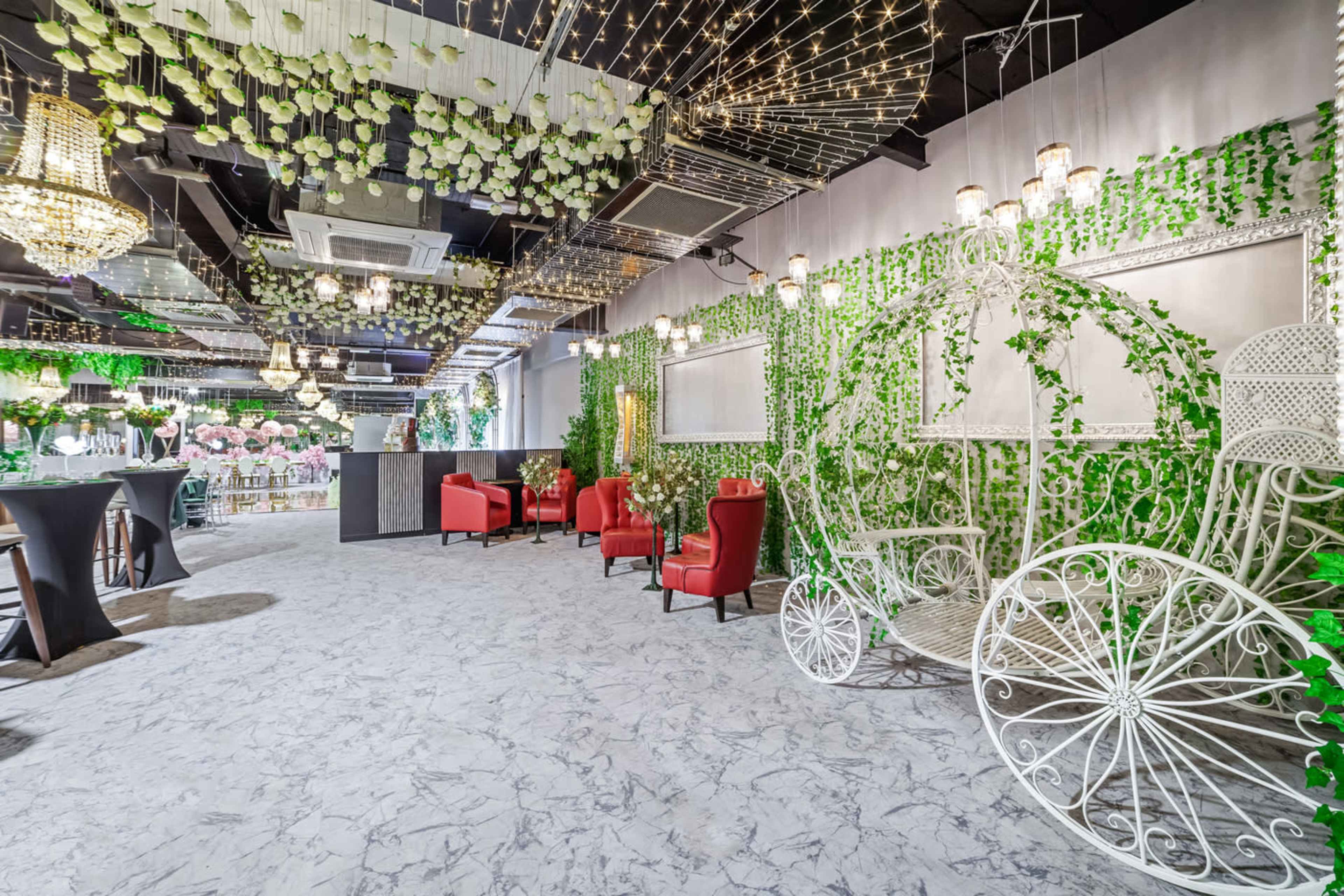 The scene shows a decorated indoor venue with green foliage, chandeliers, and red seating arranged around a whimsical white carriage structure.