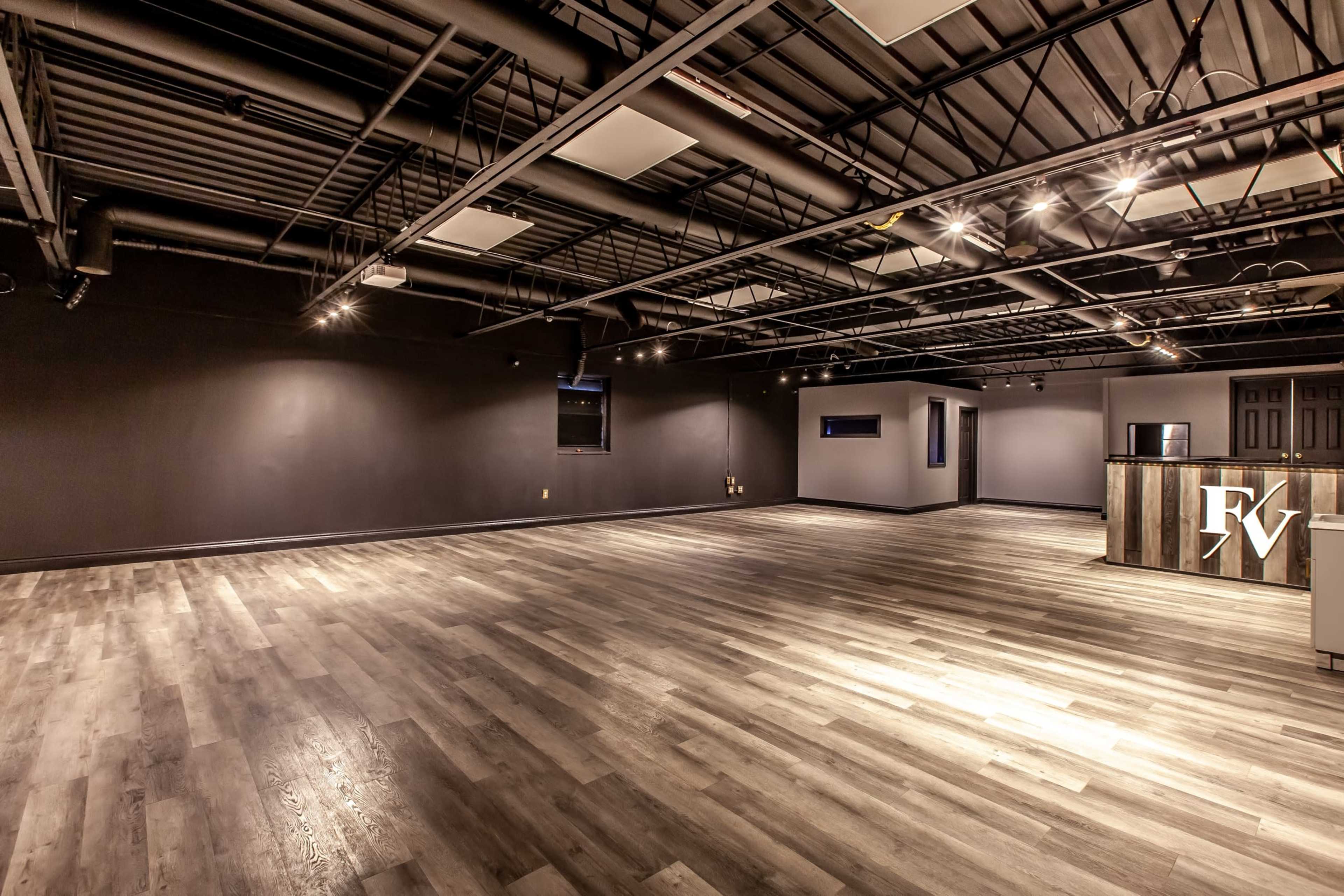 A spacious, empty room with dark walls and a wooden counter at one end, illuminated by track lighting overhead.
