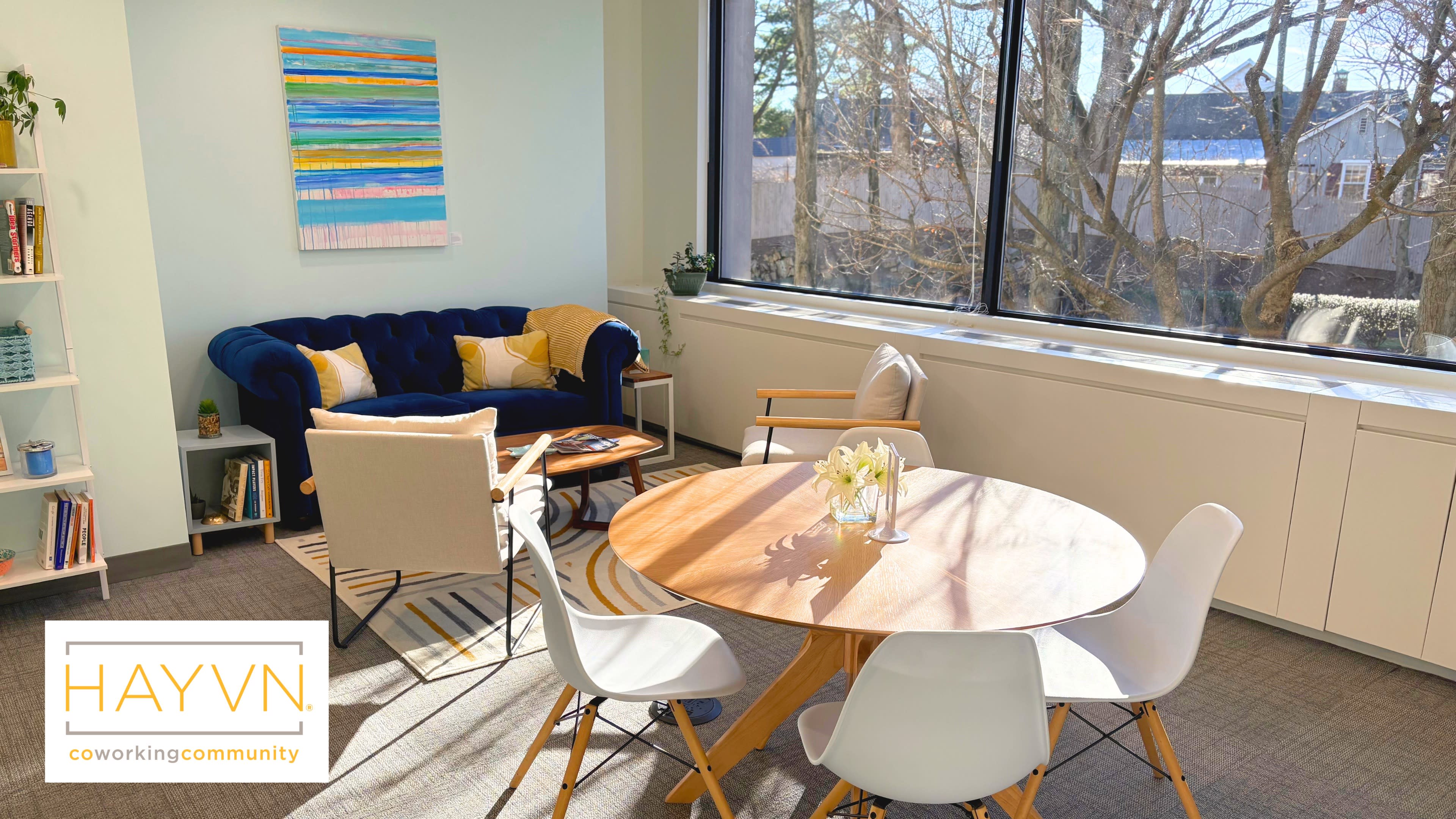 The scene shows a bright coworking space featuring a blue velvet couch, a circular wooden table surrounded by white chairs, and a large window with tree branches visible outside.