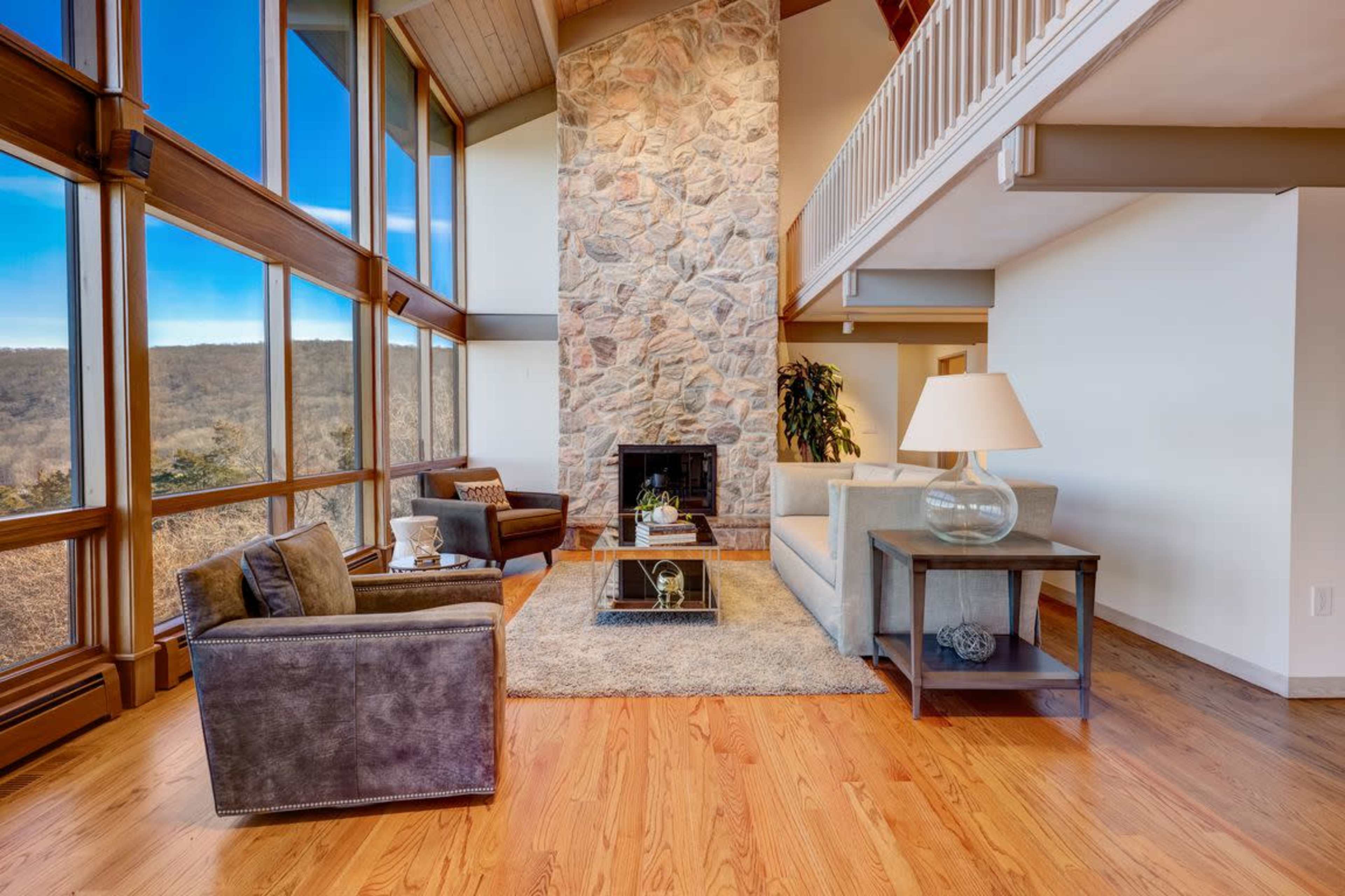 A modern living room features a large stone fireplace, large windows that offer a view of the outdoors, and a mix of comfortable seating arrangements.
