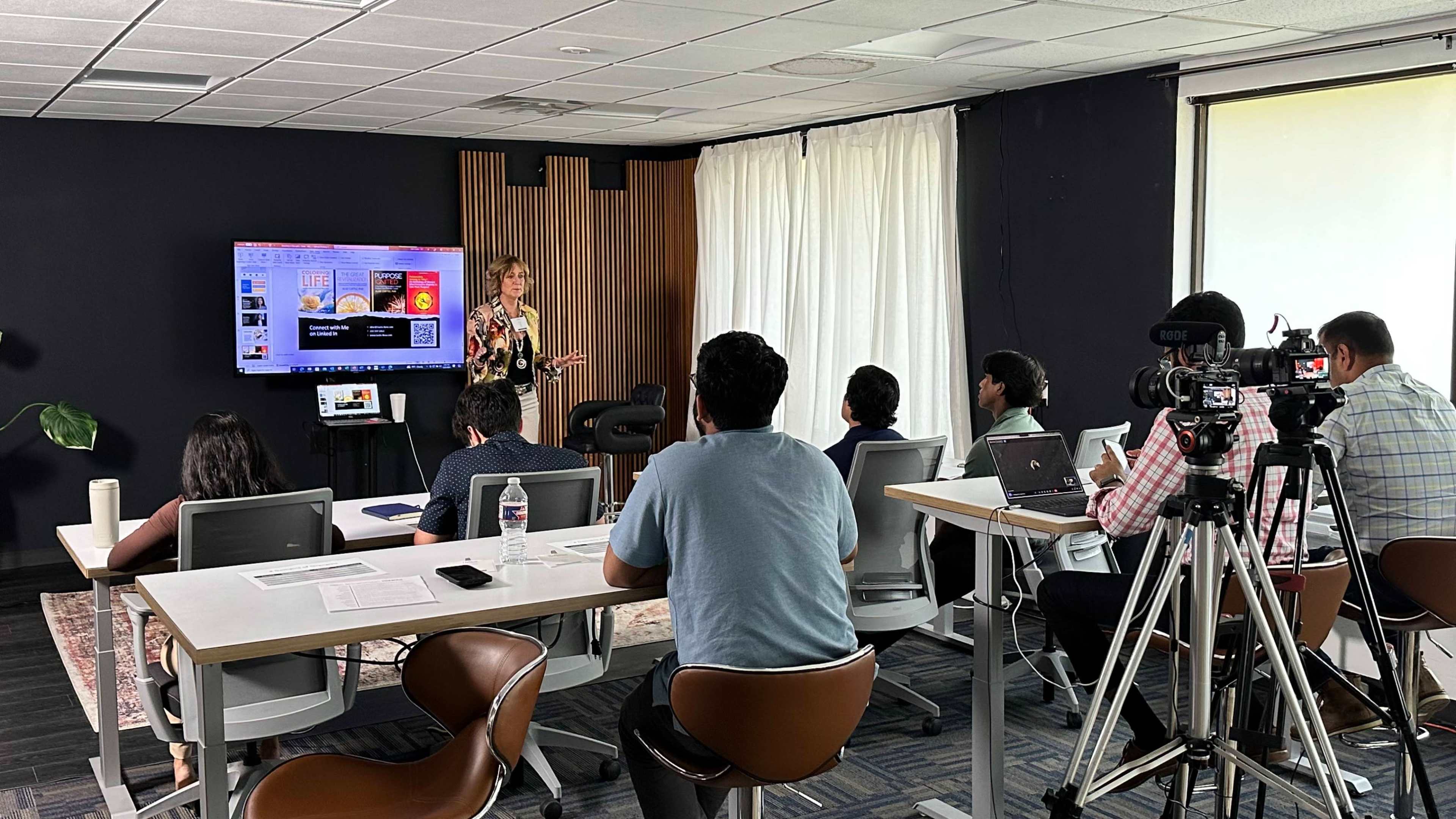 A woman stands at the front of a modern classroom, presenting to a group of seated students while a camera captures the session.
