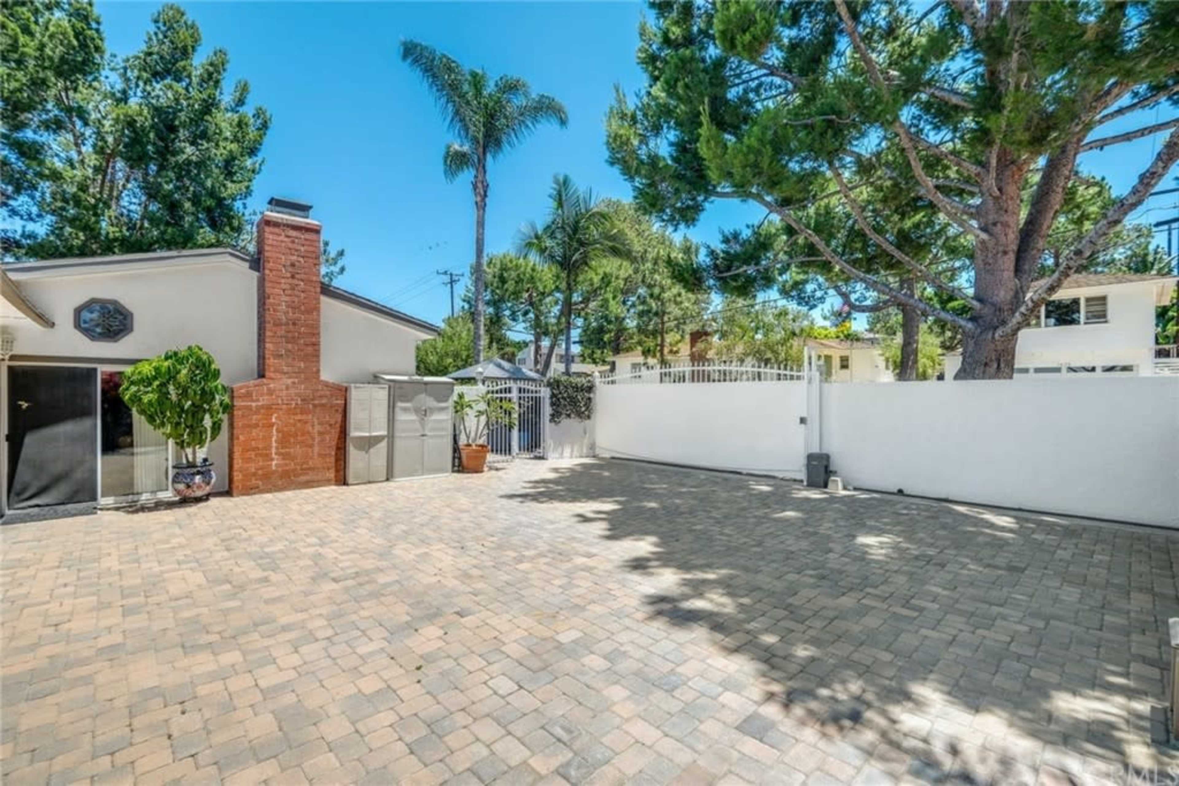 The image shows a paved outdoor space bordered by trees and a low white wall, with a house and a chimney in the background.