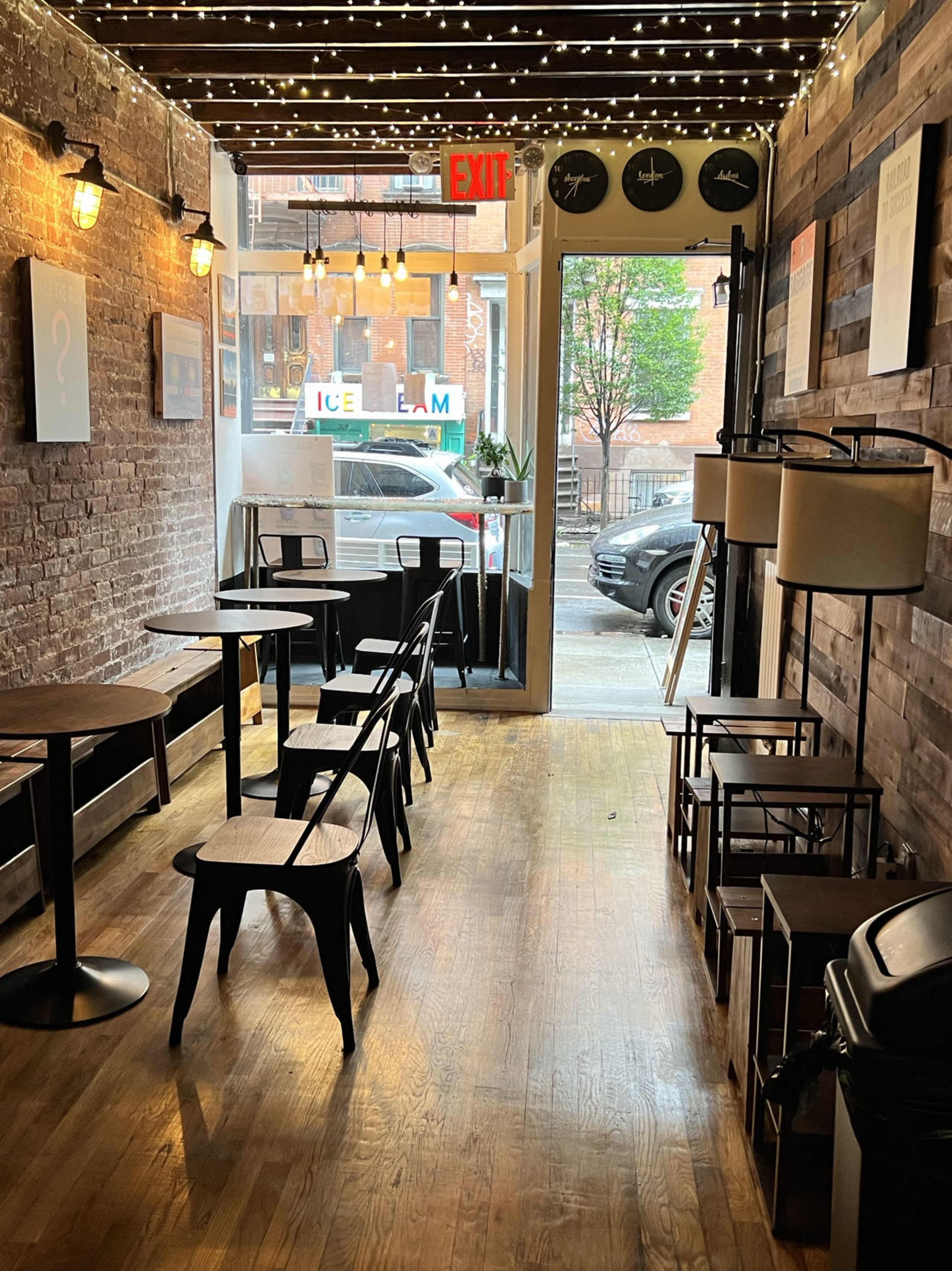 A narrow café with wooden flooring and exposed brick walls, featuring several tables and chairs arranged along the length of the space, leading to a doorway with an exit sign.