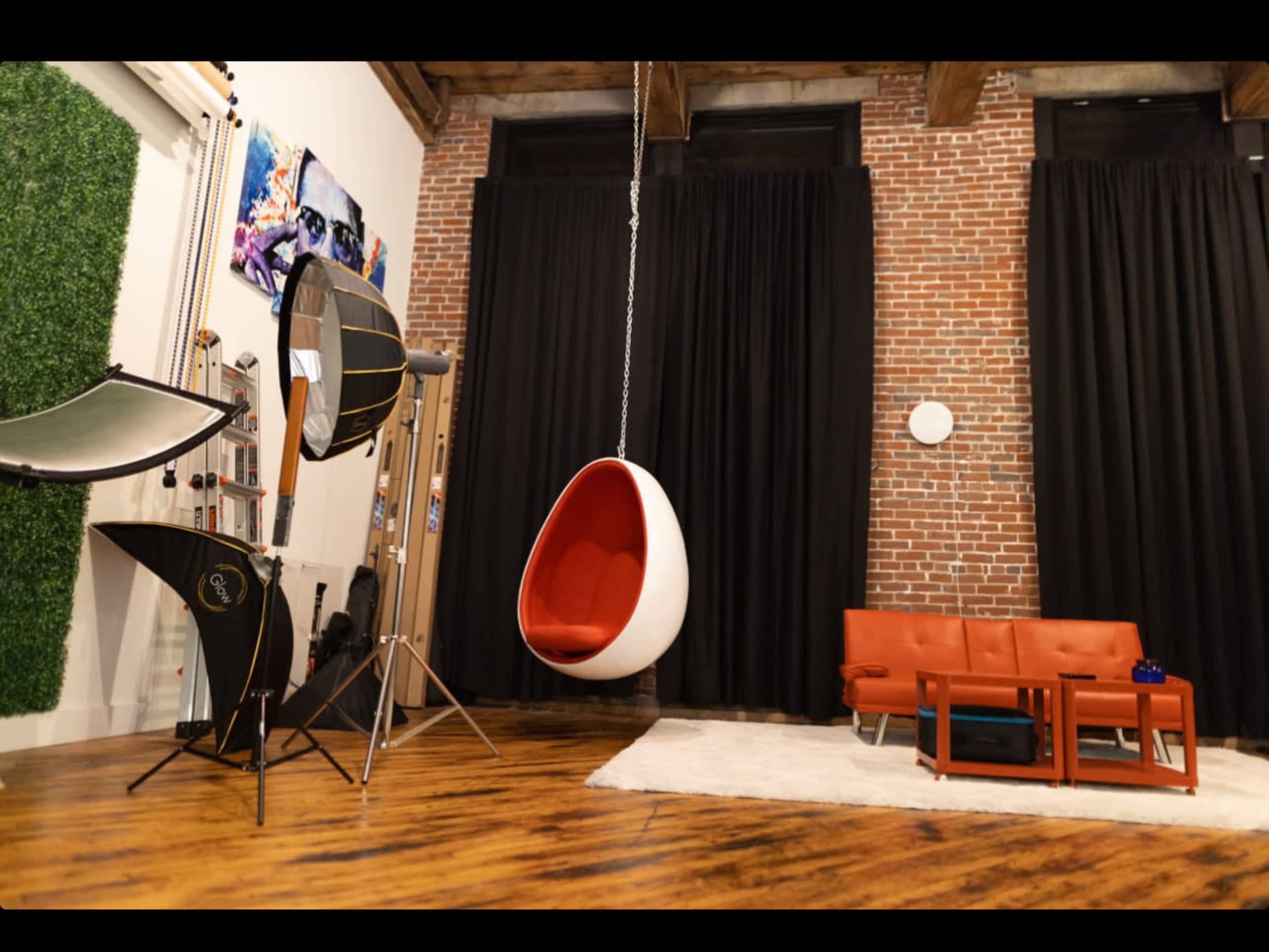 A modern living space features a hanging white egg chair, a red leather sofa, a wooden coffee table, and a backdrop of exposed brick walls.