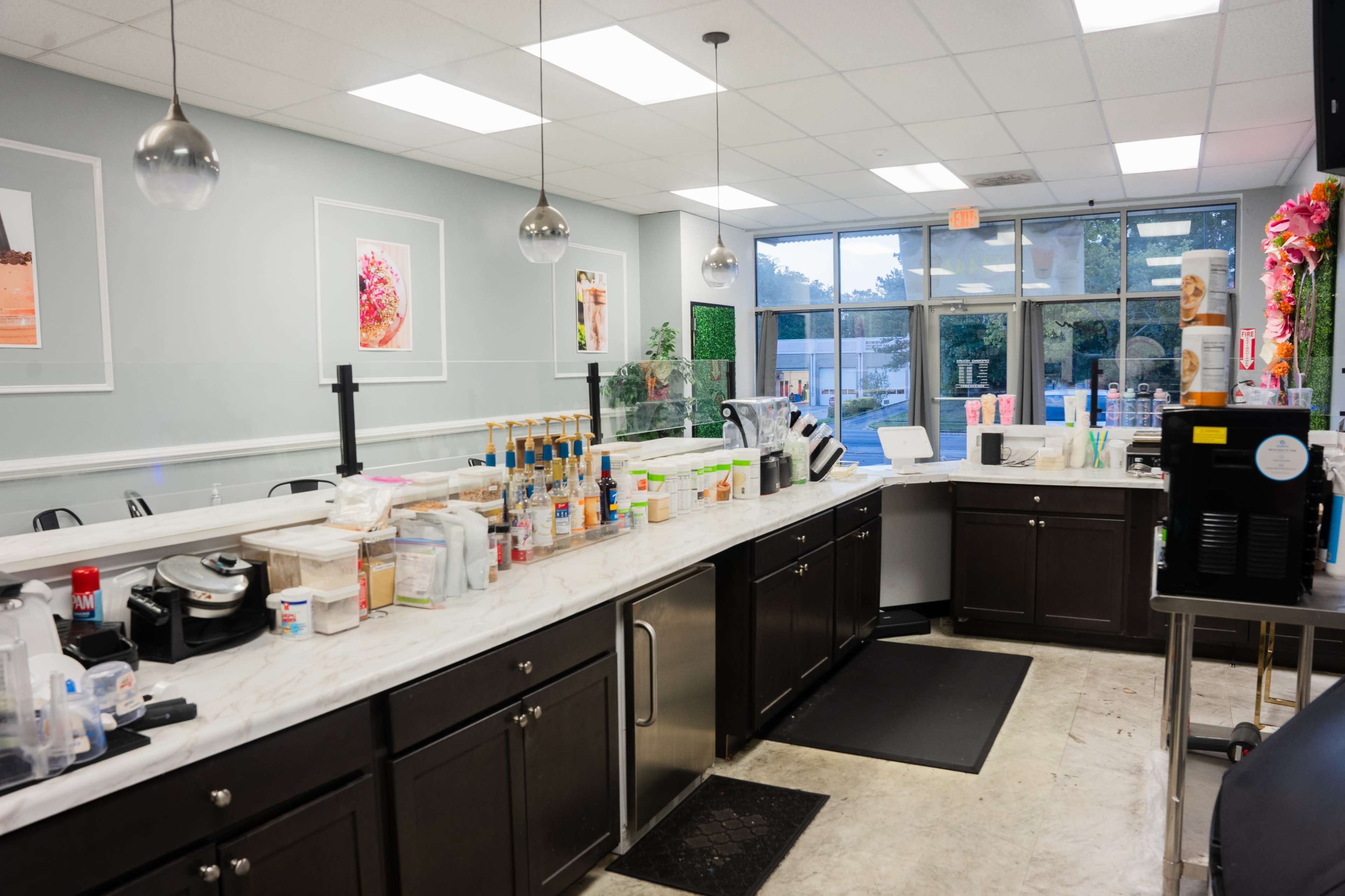 The image shows a modern cafe kitchen with a marble countertop, various beverage containers, a sink, and large windows letting in natural light.