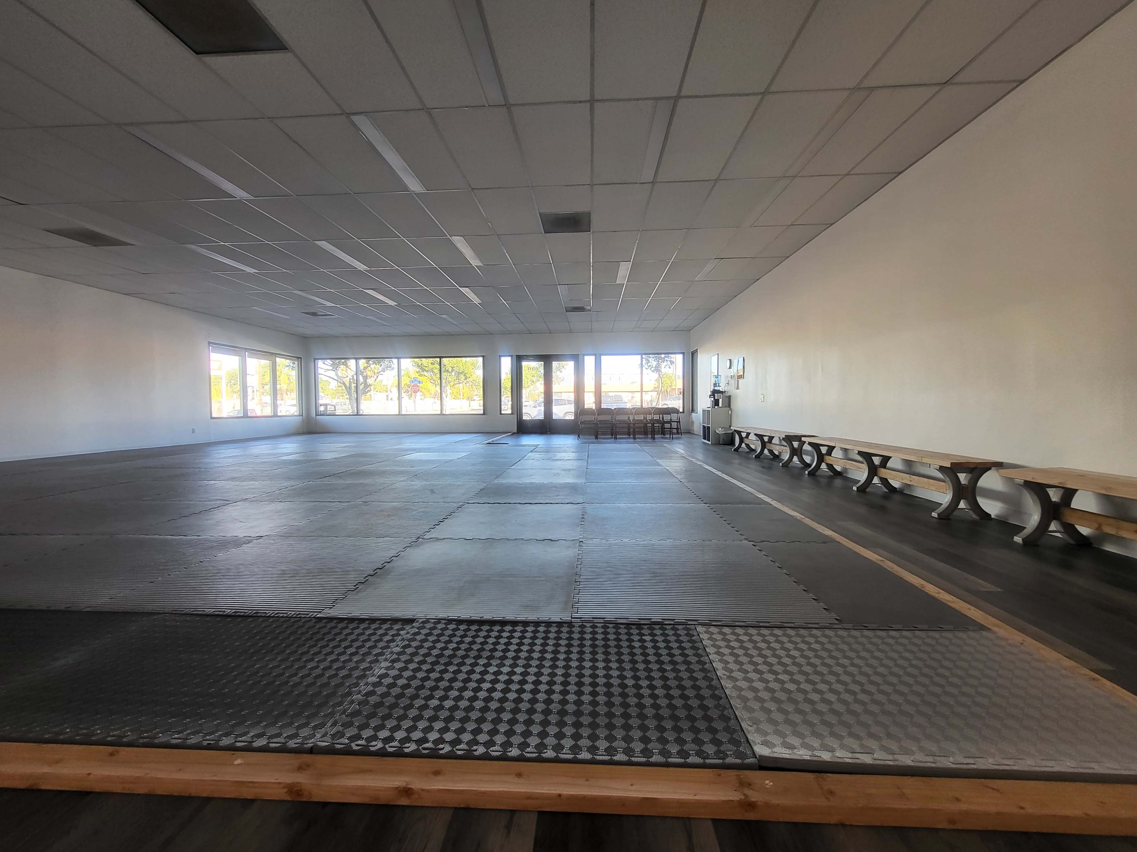 The image shows a spacious, empty room with a padded flooring area and benches along one wall, featuring large windows that let in sunlight.