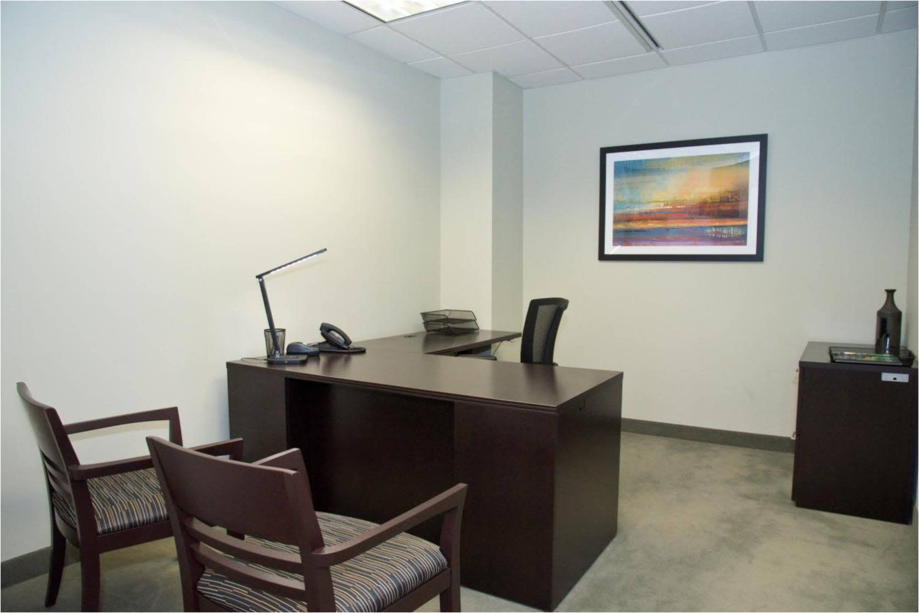 The image shows a minimalistic office space featuring a desk, an office chair, two visitor chairs, a phone, and a wall-mounted artwork.