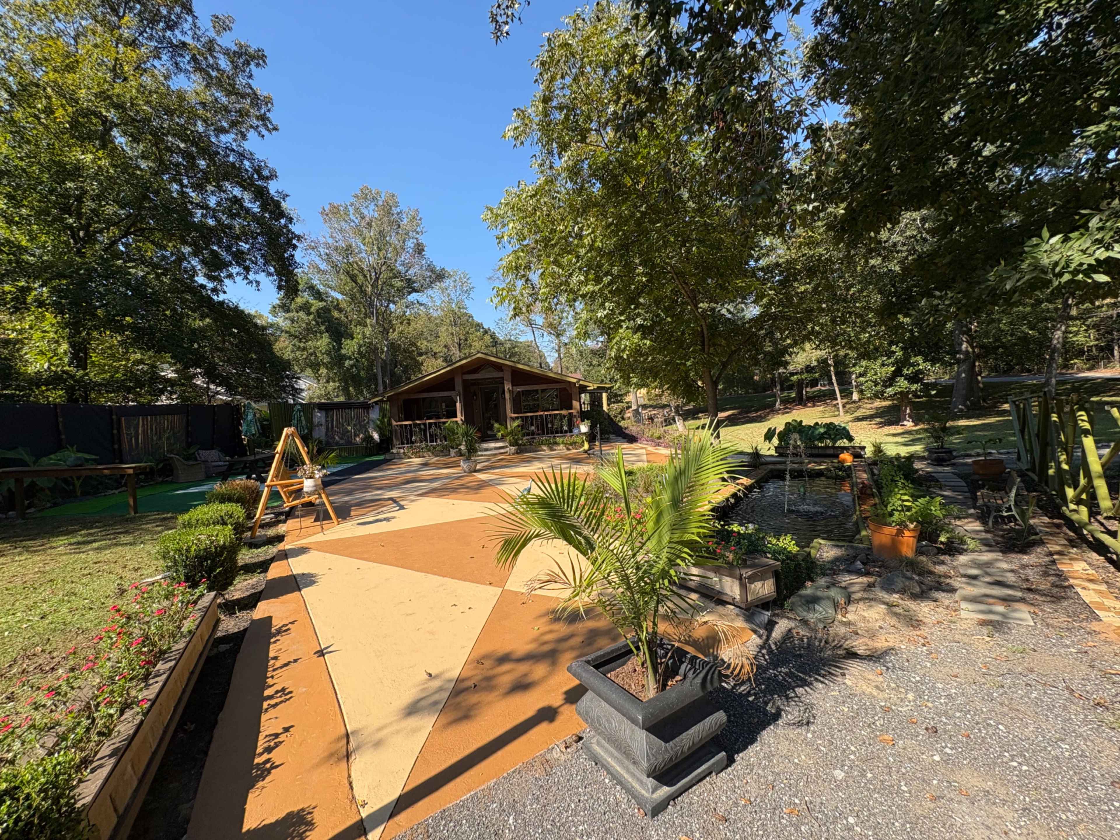 The image shows a landscaped outdoor area featuring a patio with orange and cream-colored sections, surrounded by greenery and various planters.