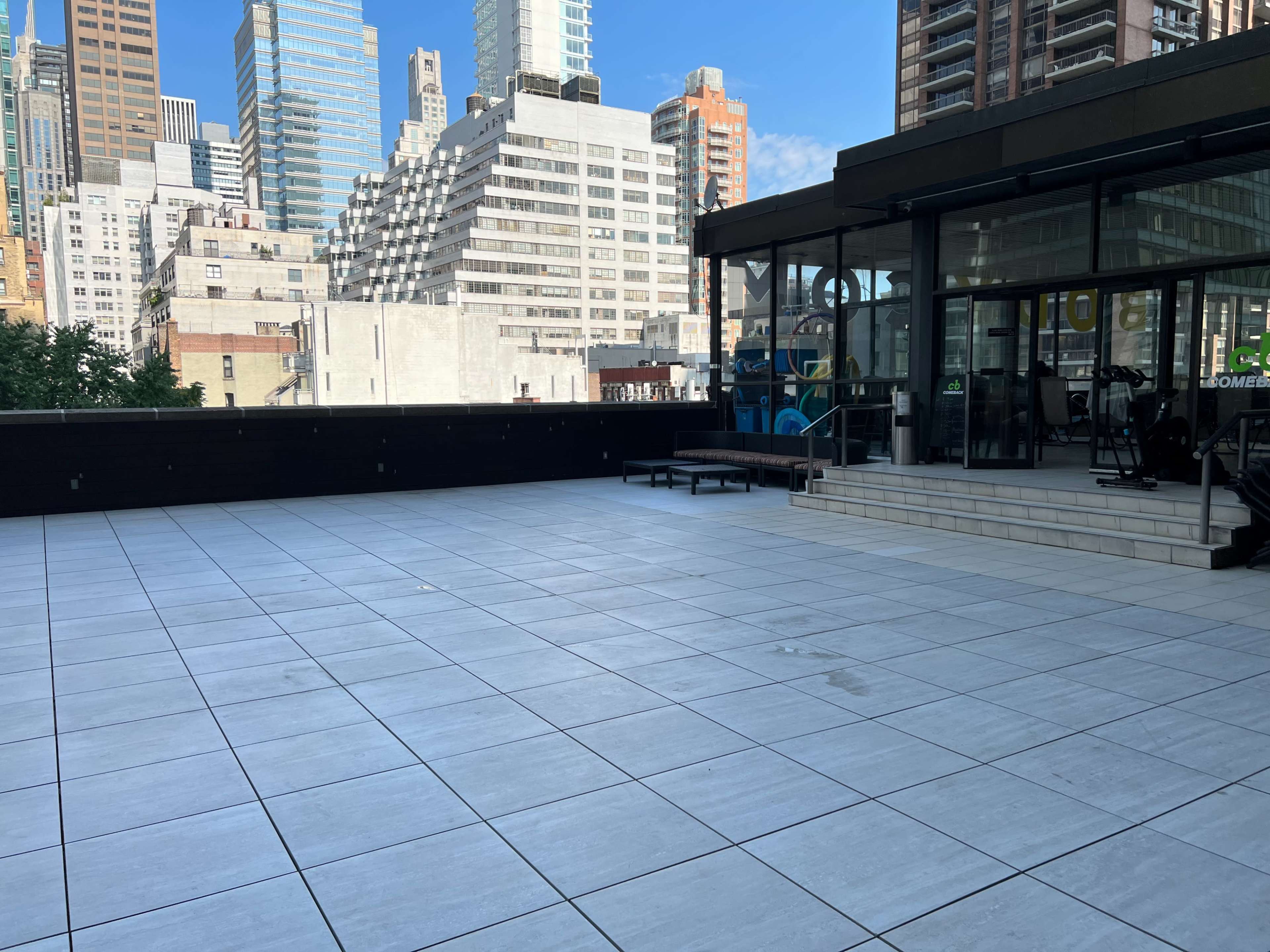 The image shows a spacious outdoor terrace with large gray tiles, surrounded by tall city buildings.