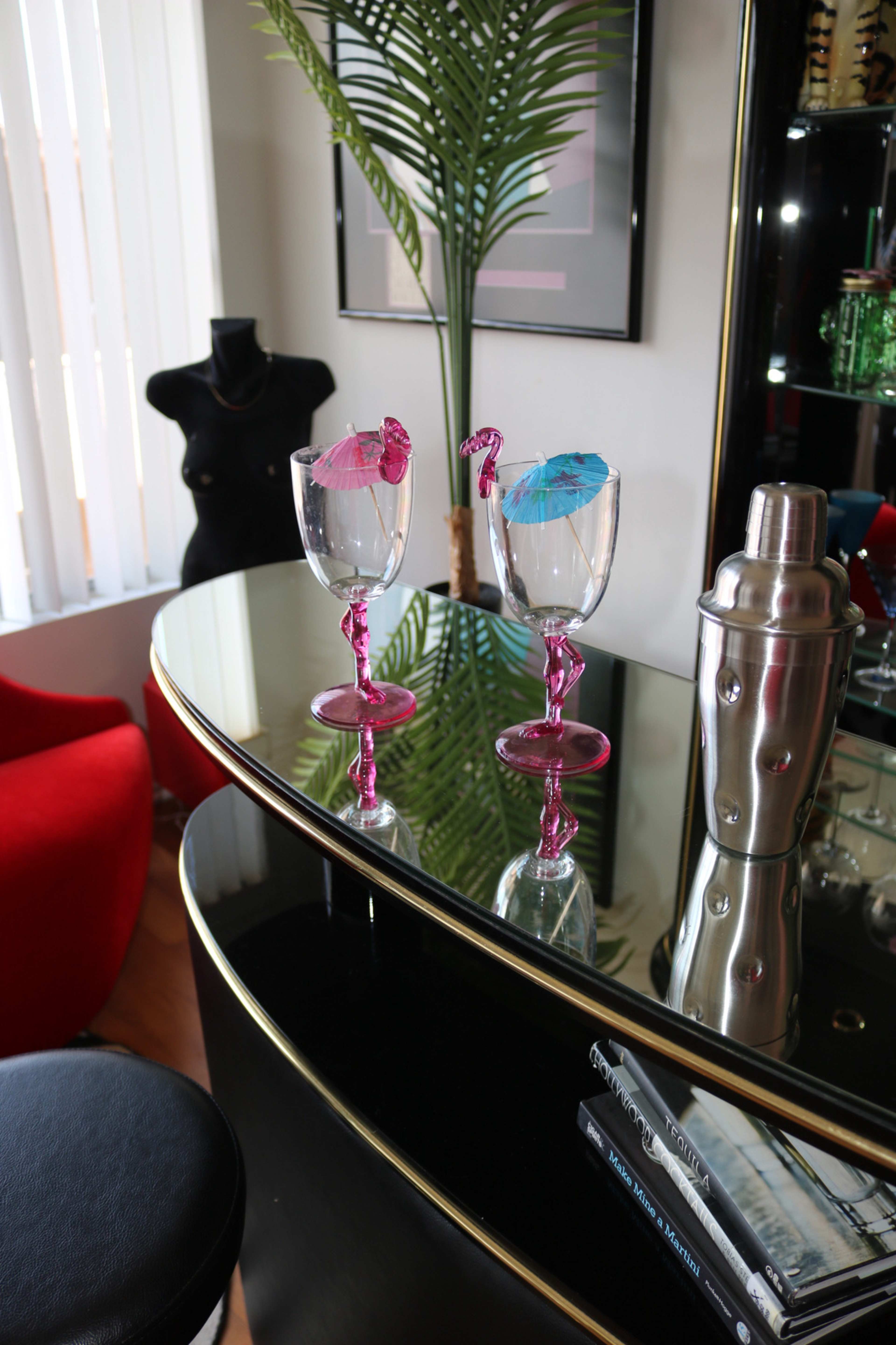 Two stemmed glasses with colorful umbrellas sit on a reflective black bar countertop next to a palm plant and a red chair.