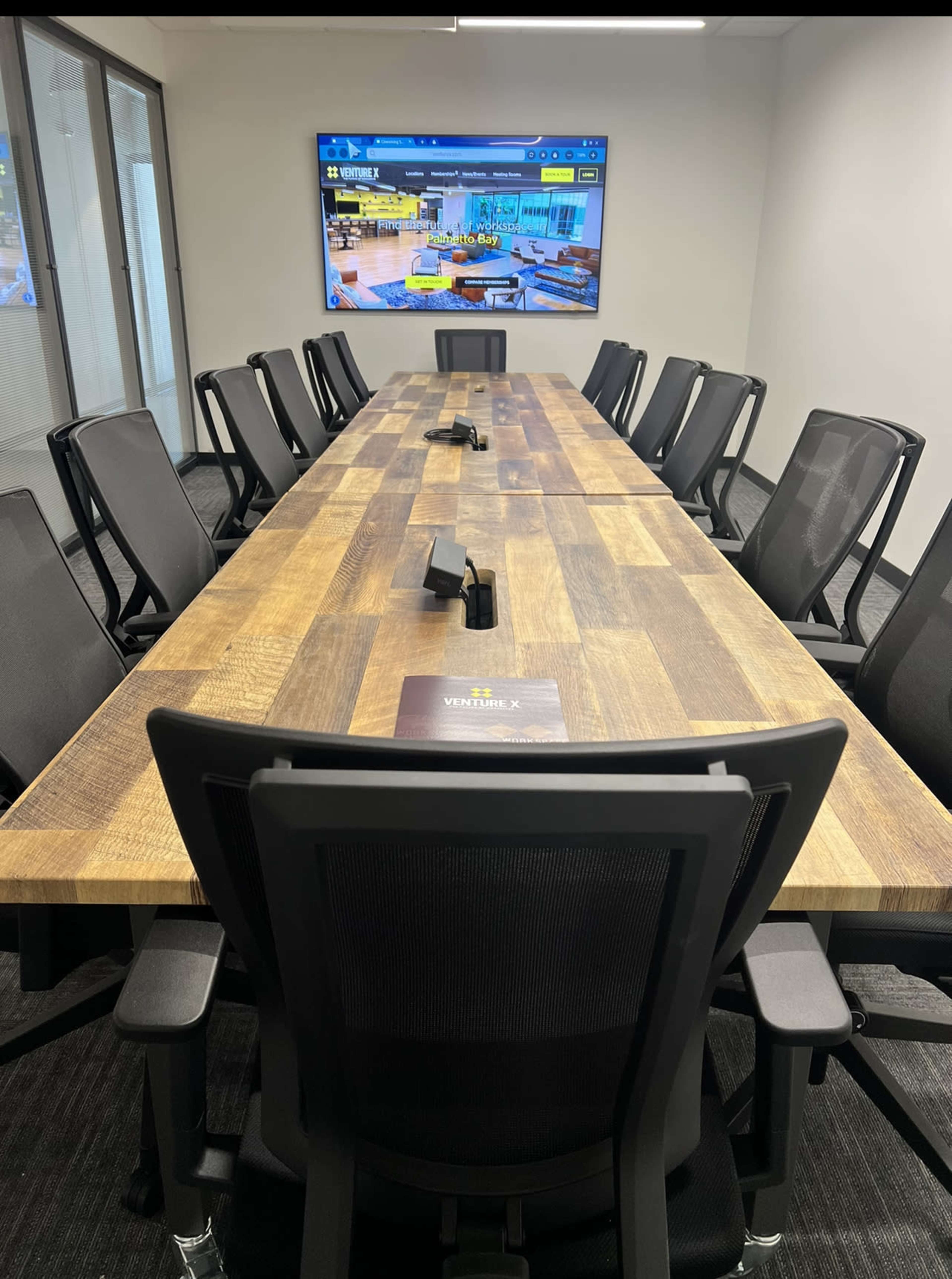 A long wooden conference table with multiple chairs is set up in a room featuring a large screen display on the wall.
