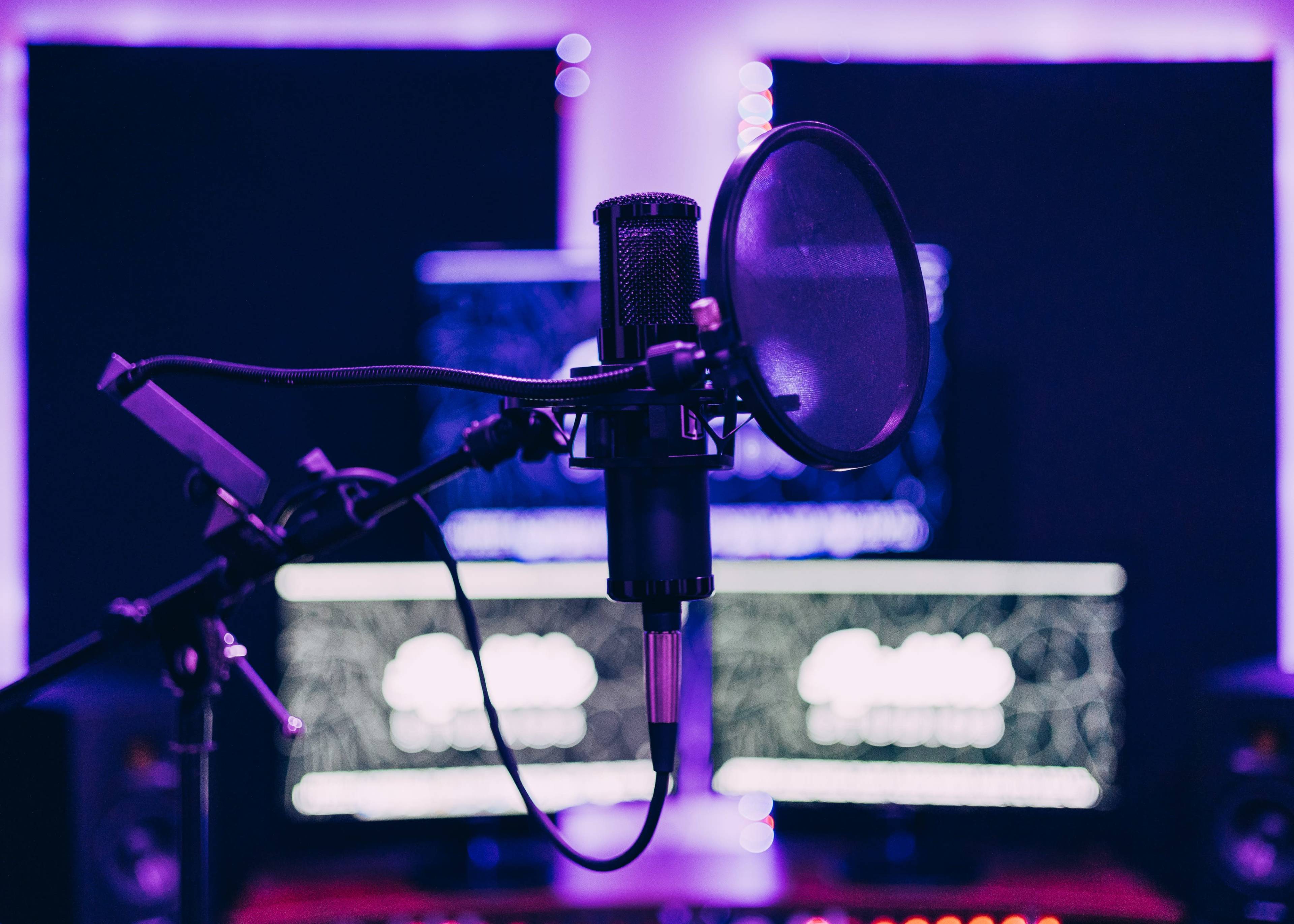 A microphone on a stand is positioned in front of two computer screens in a sound studio.
