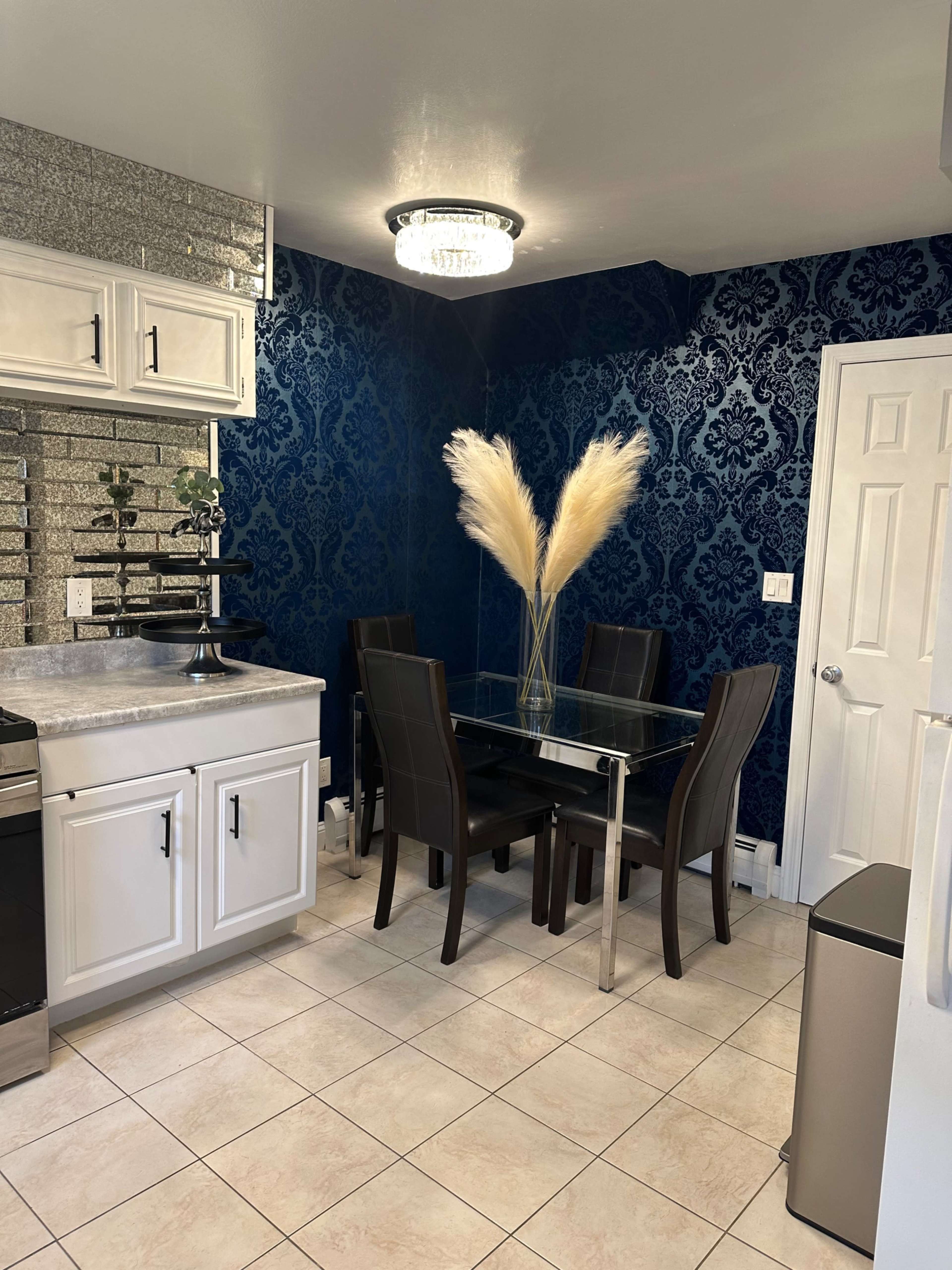 A modern dining area features a glass table surrounded by black chairs, set against a blue patterned wall and illuminated by a chandelier.