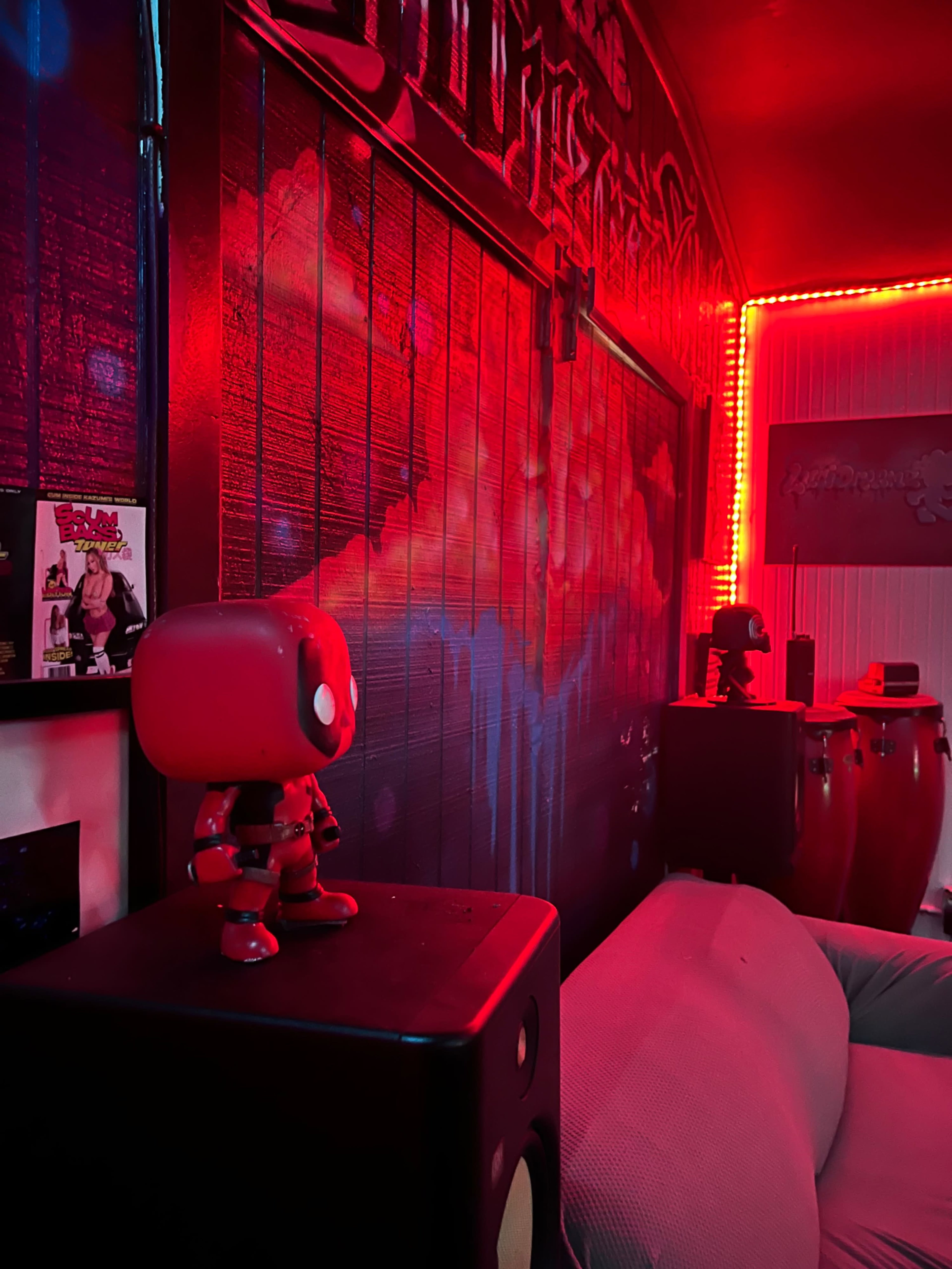The image shows a corner of a room featuring a Deadpool figurine on a speaker, with colorful wall art and ambient red lighting.