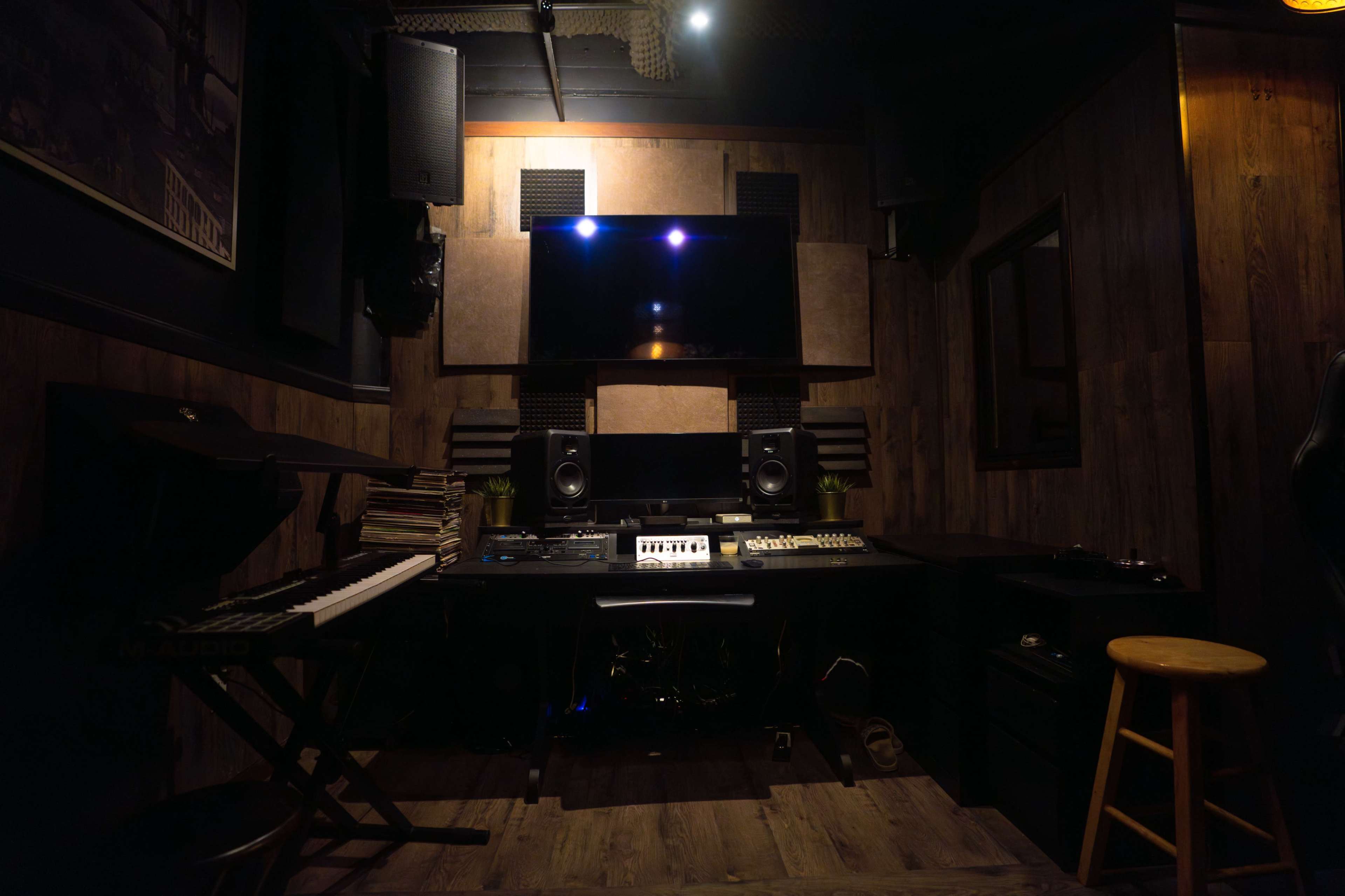 A music studio features a wooden interior, a keyboard on the left, speakers mounted on the walls, and a central console with audio equipment.