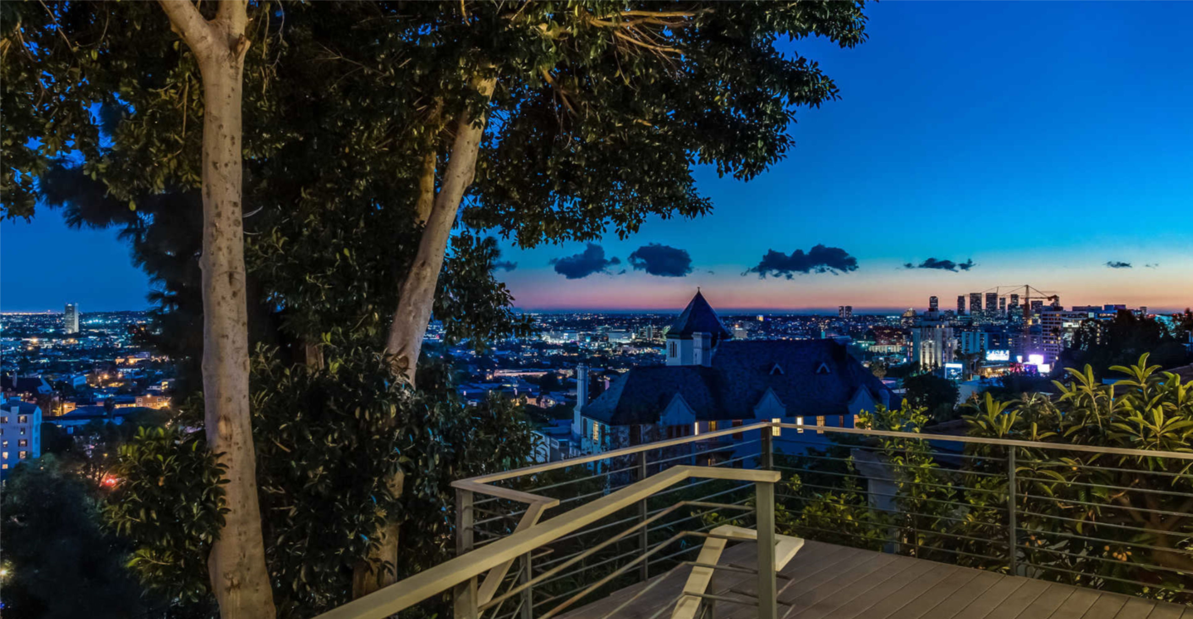 The image shows a city skyline at dusk with a terrace and trees in the foreground.