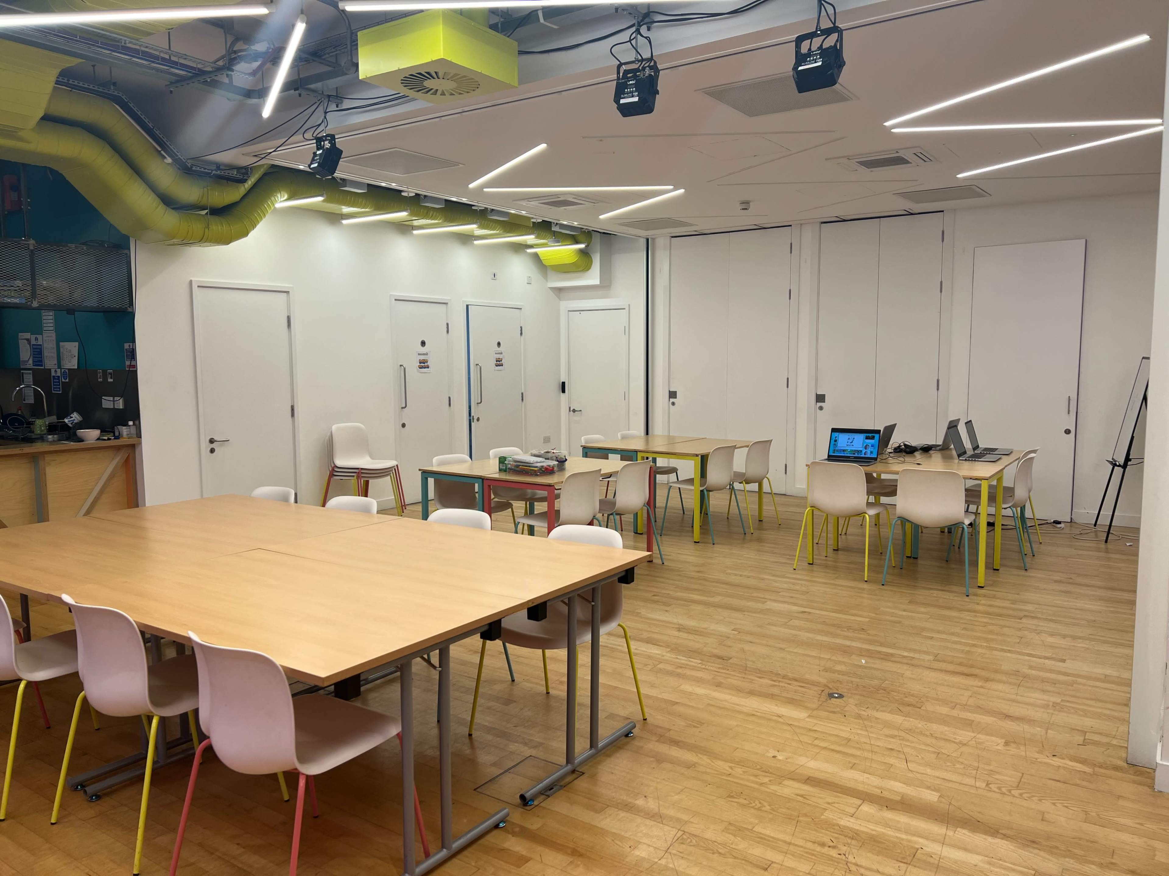 A brightly lit room features several wooden tables surrounded by chairs, with a laptop on one table and doors leading to separate spaces.