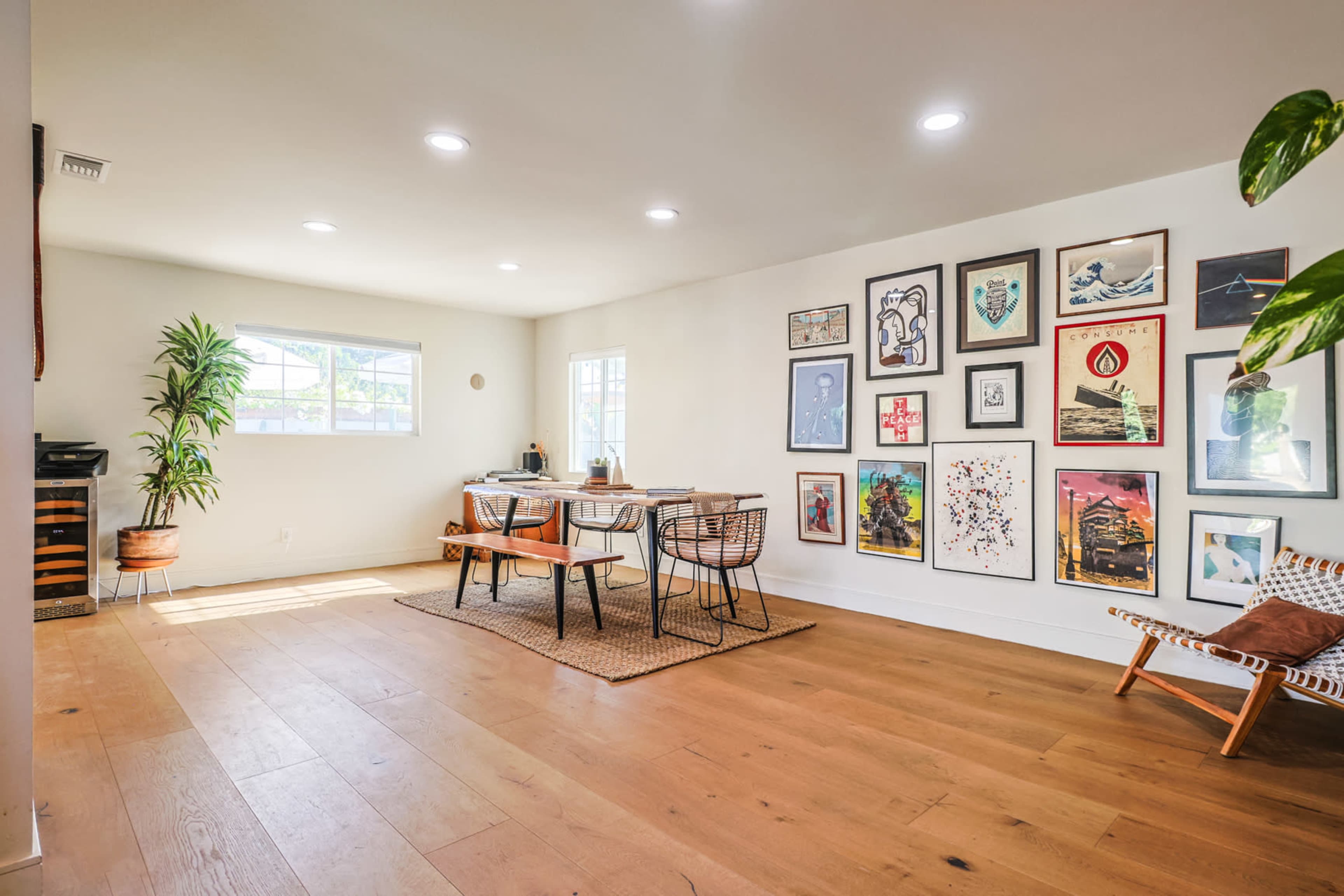 A bright room with a dining table, chairs, various framed artworks on the walls, and a potted plant in one corner.