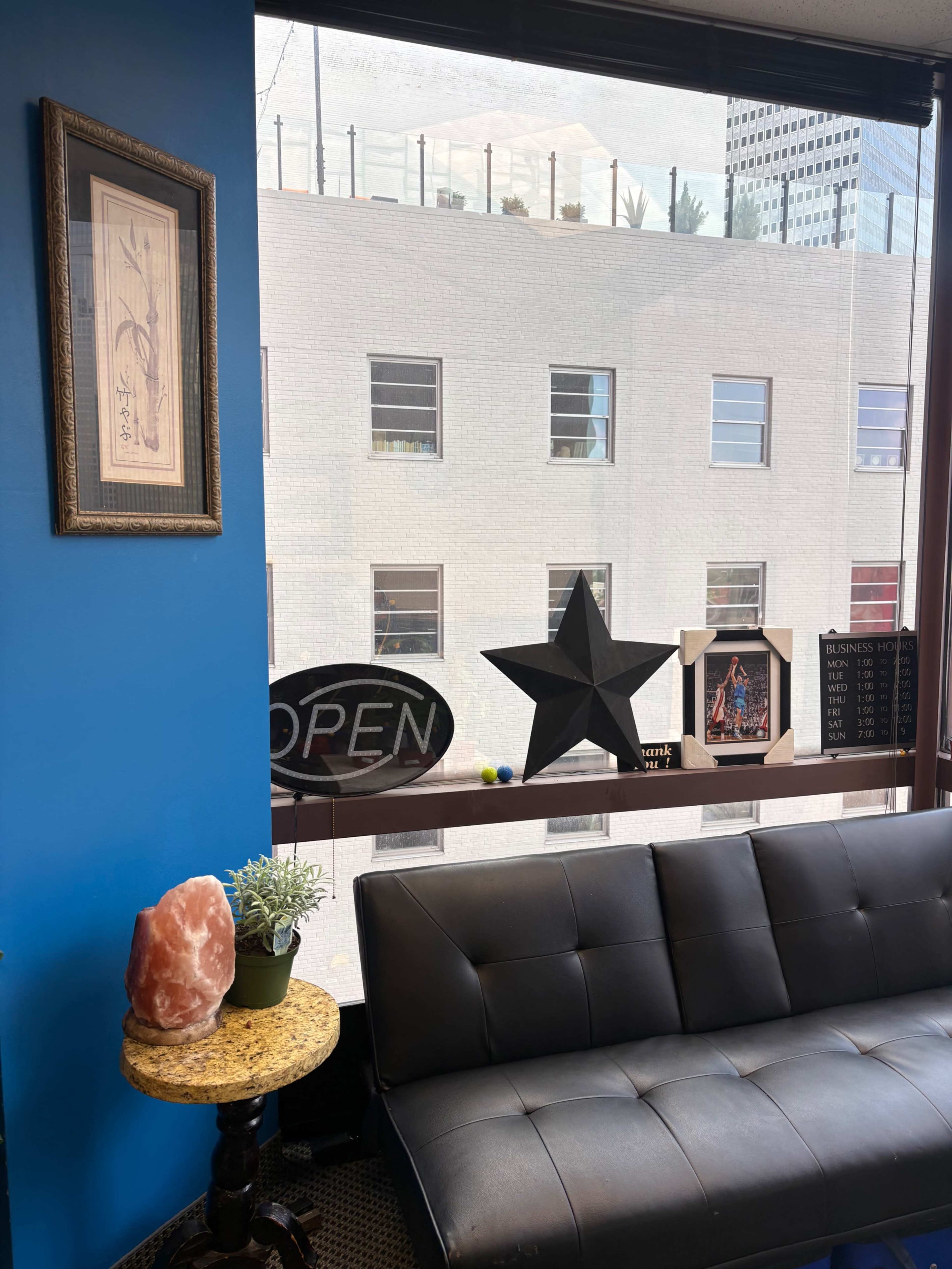 The image shows a cozy corner of a room with a black couch, a small round table featuring a plant and a salt lamp, and a window displaying a cityscape with various decorative items on the windowsill.