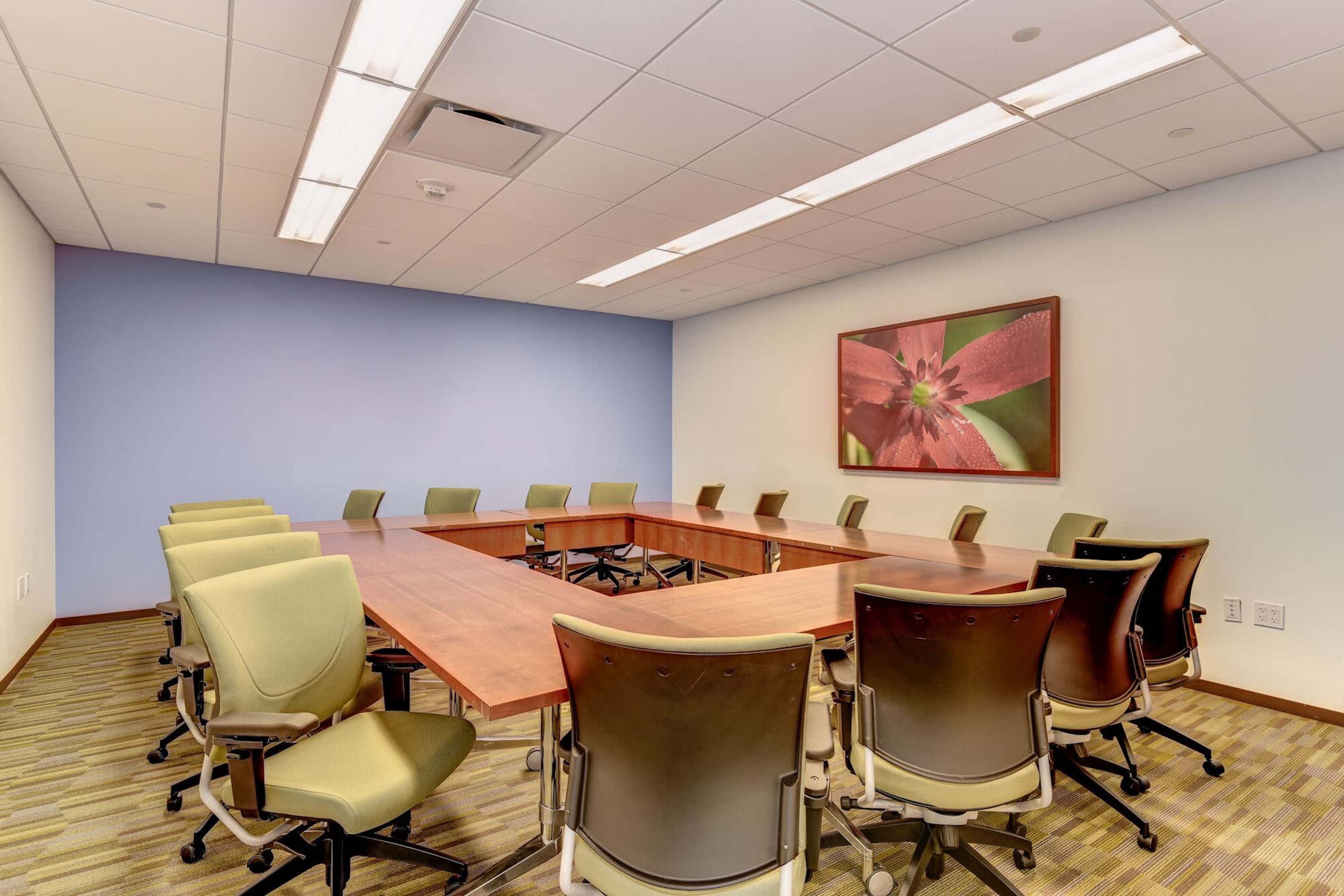 A conference room features a large wooden table surrounded by ergonomic chairs, with a floral artwork on the wall and blue accents.