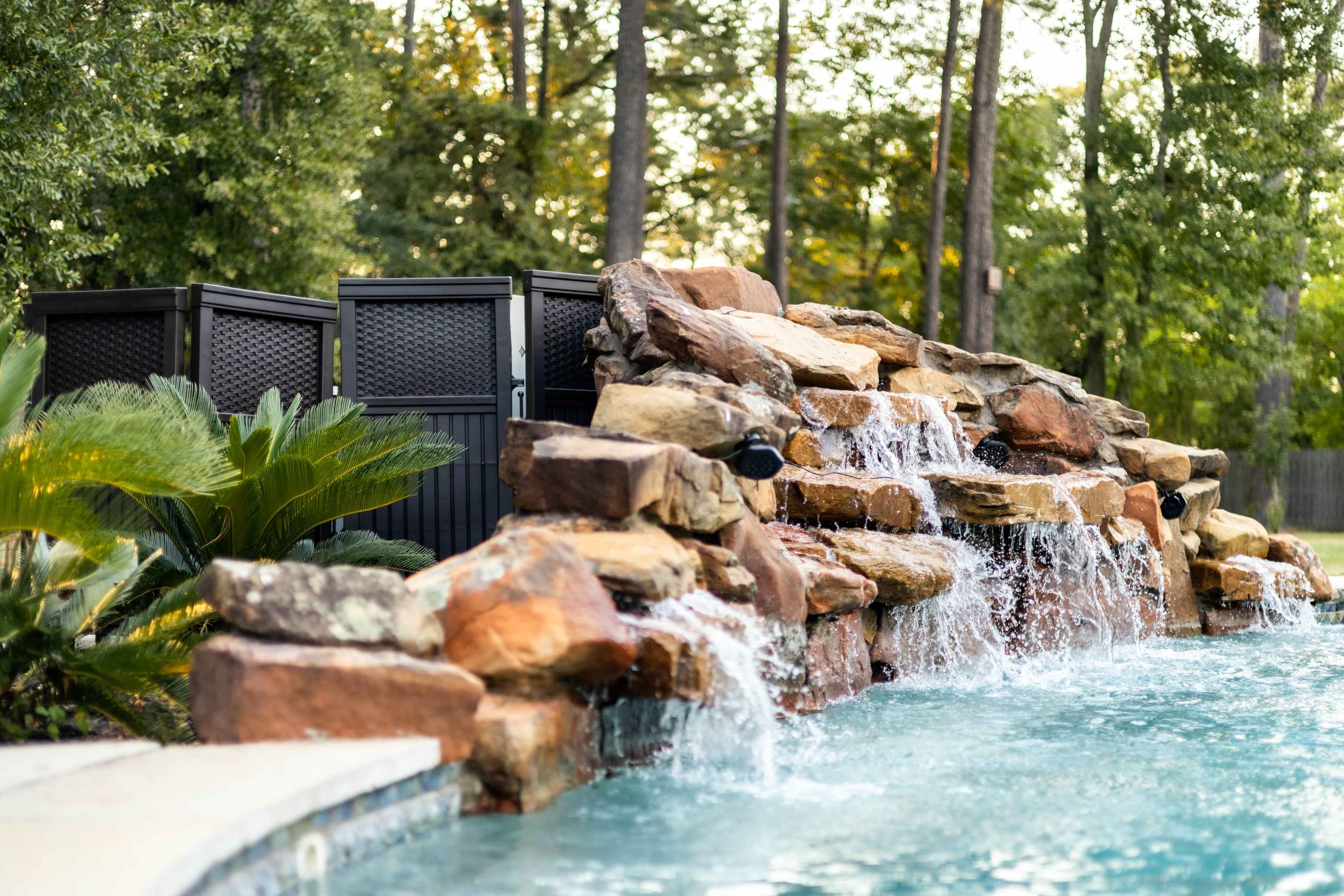 A stone waterfall cascades into a blue pool surrounded by greenery and a privacy screen in a wooded backyard.