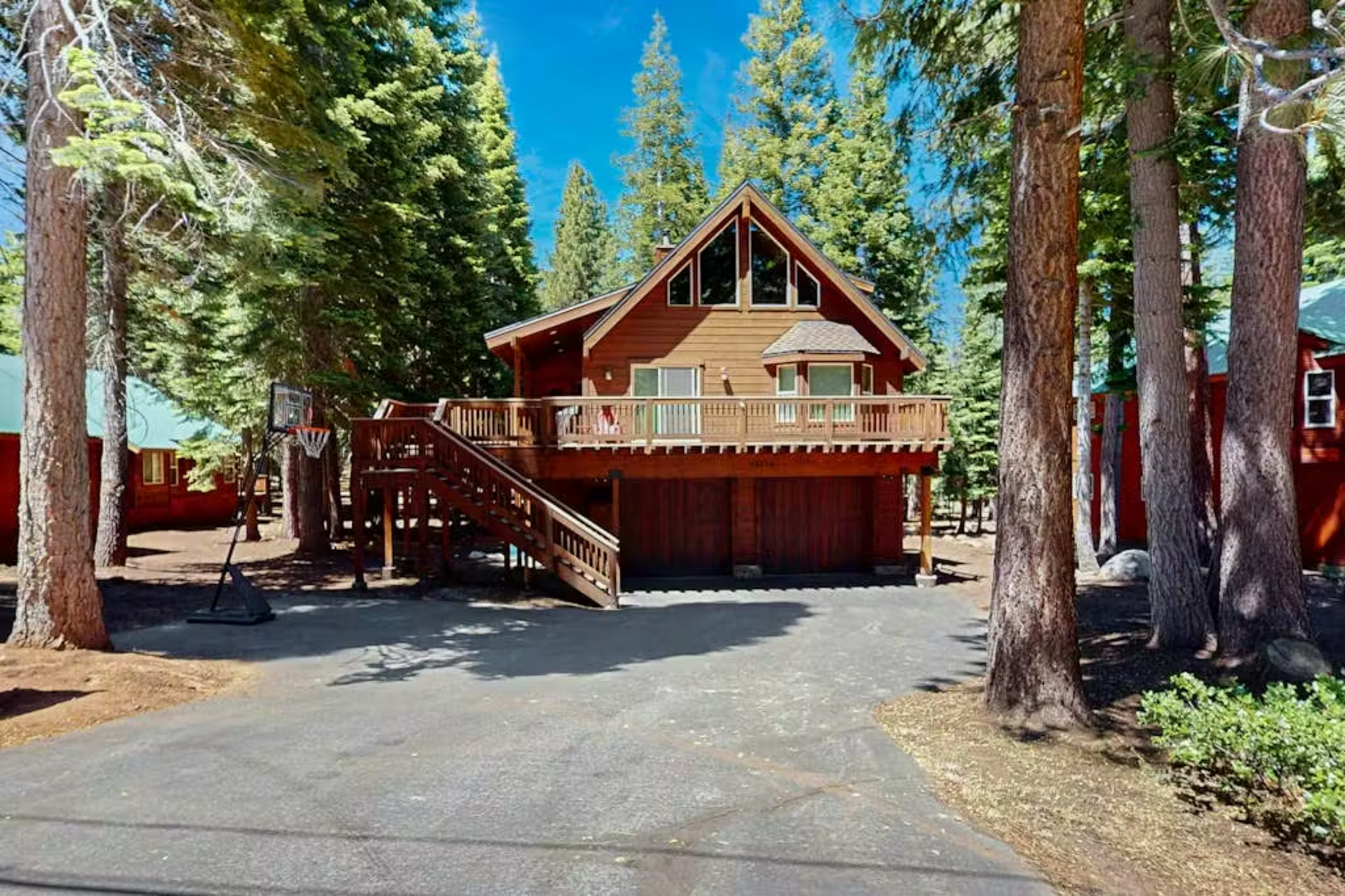A two-story brown wooden cabin with a deck is situated among tall pine trees, with a basketball hoop visible in the driveway.