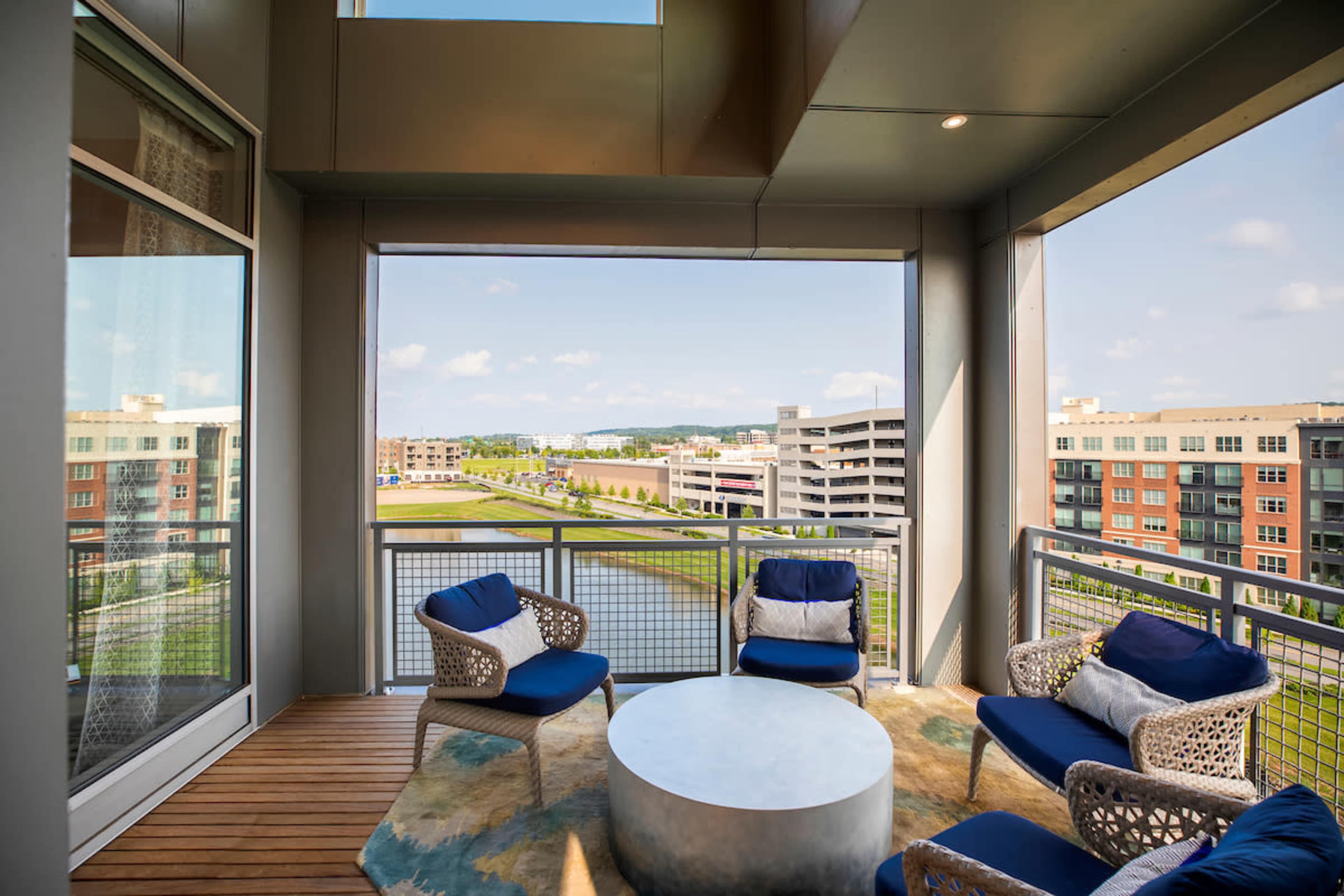 A balcony with modern seating overlooks a green area and parking structures beside a body of water.