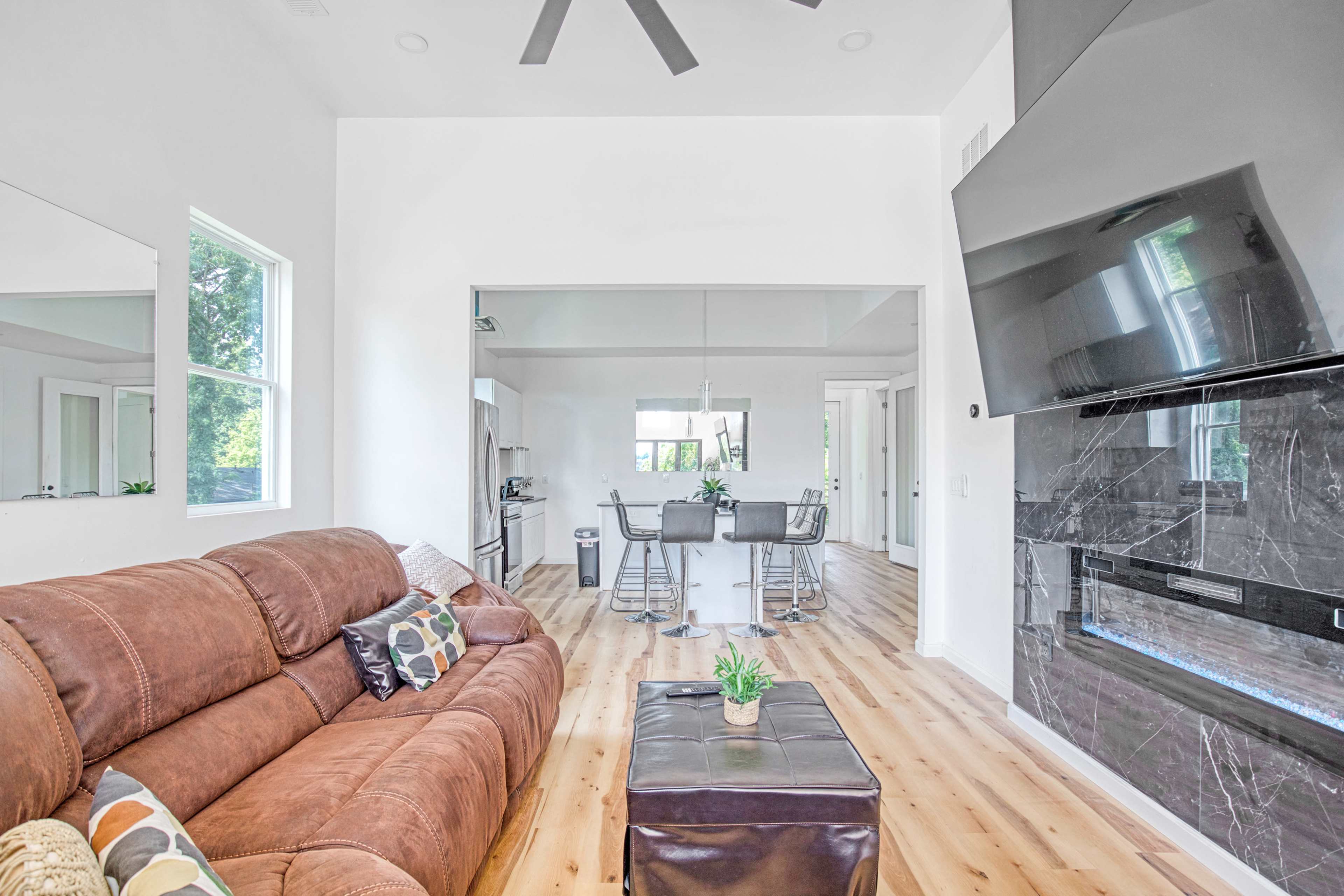 The image shows a modern living area with a brown sectional sofa, a coffee table, and an open view to a dining area and kitchen featuring a bar with stools.