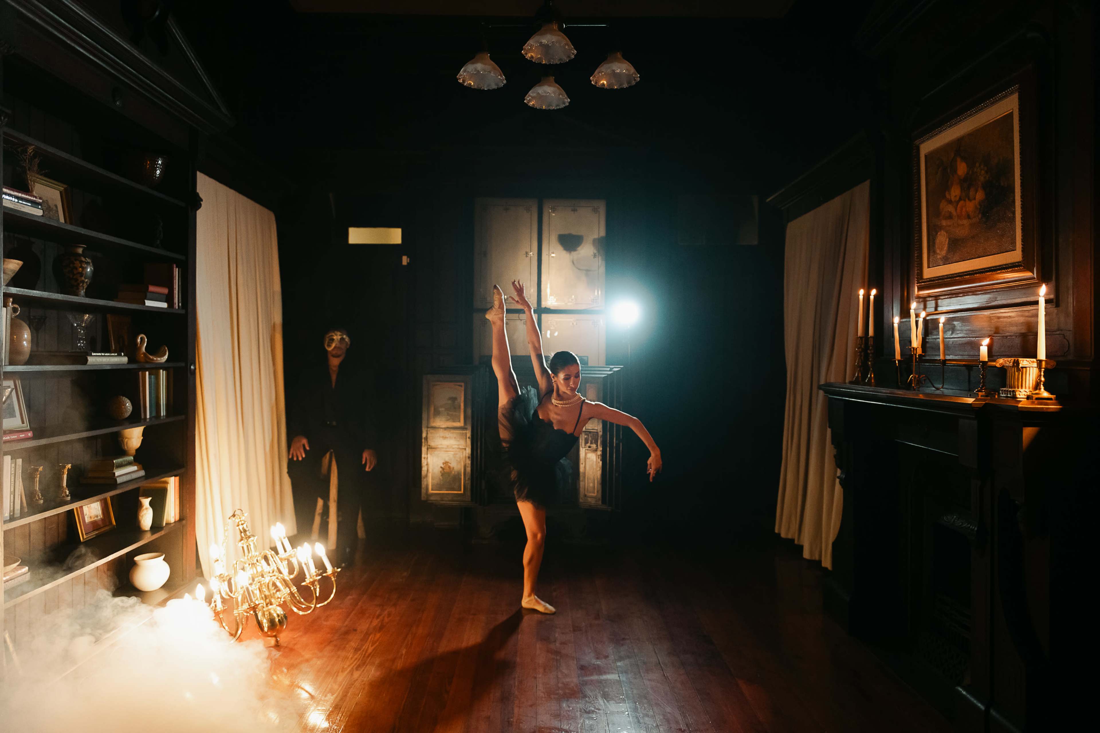 A ballerina performs an elegant pose on one leg in a dimly lit room with a chandelier and an ominous figure in the background.