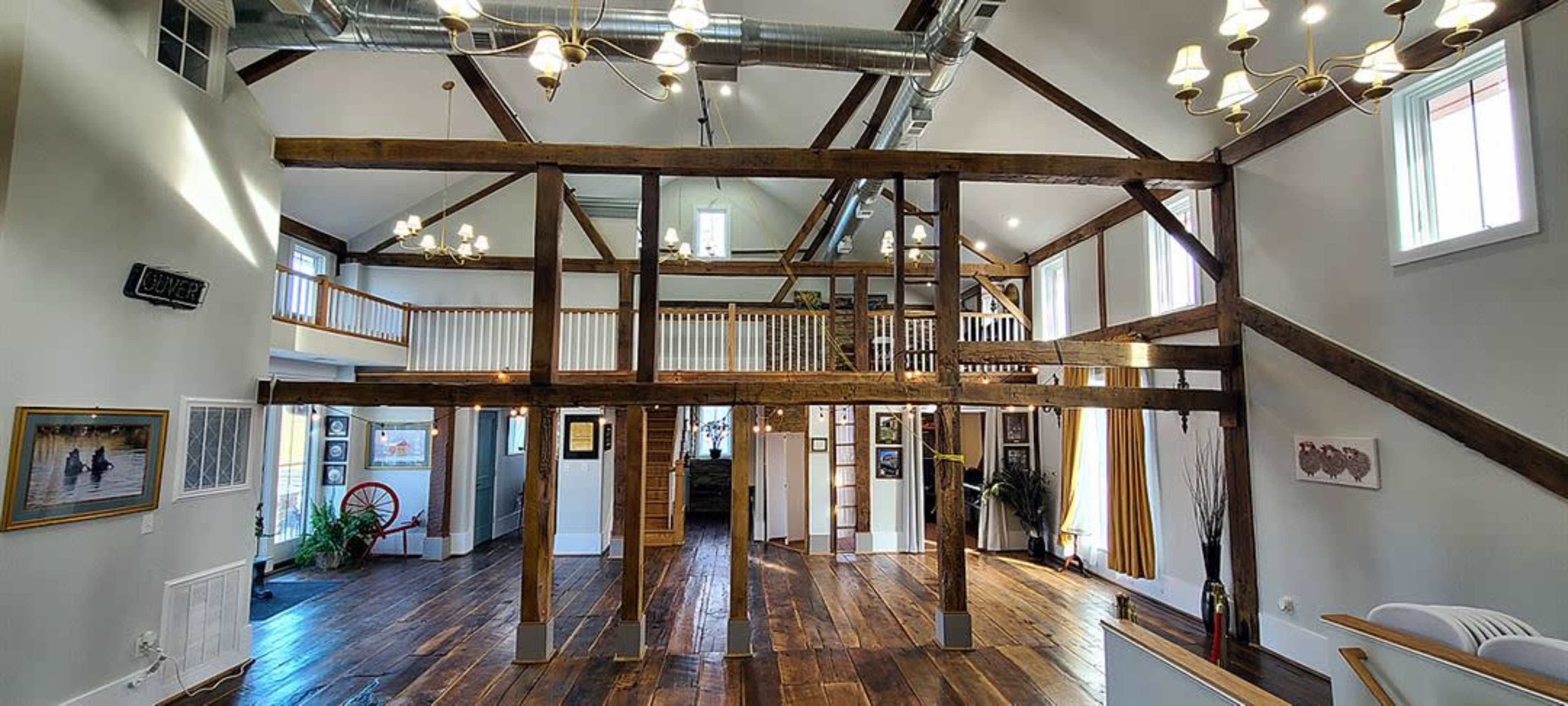The image shows the interior of a spacious room with wooden beams, high ceilings, and a welcoming atmosphere, featuring a mix of natural light and displayed artwork.