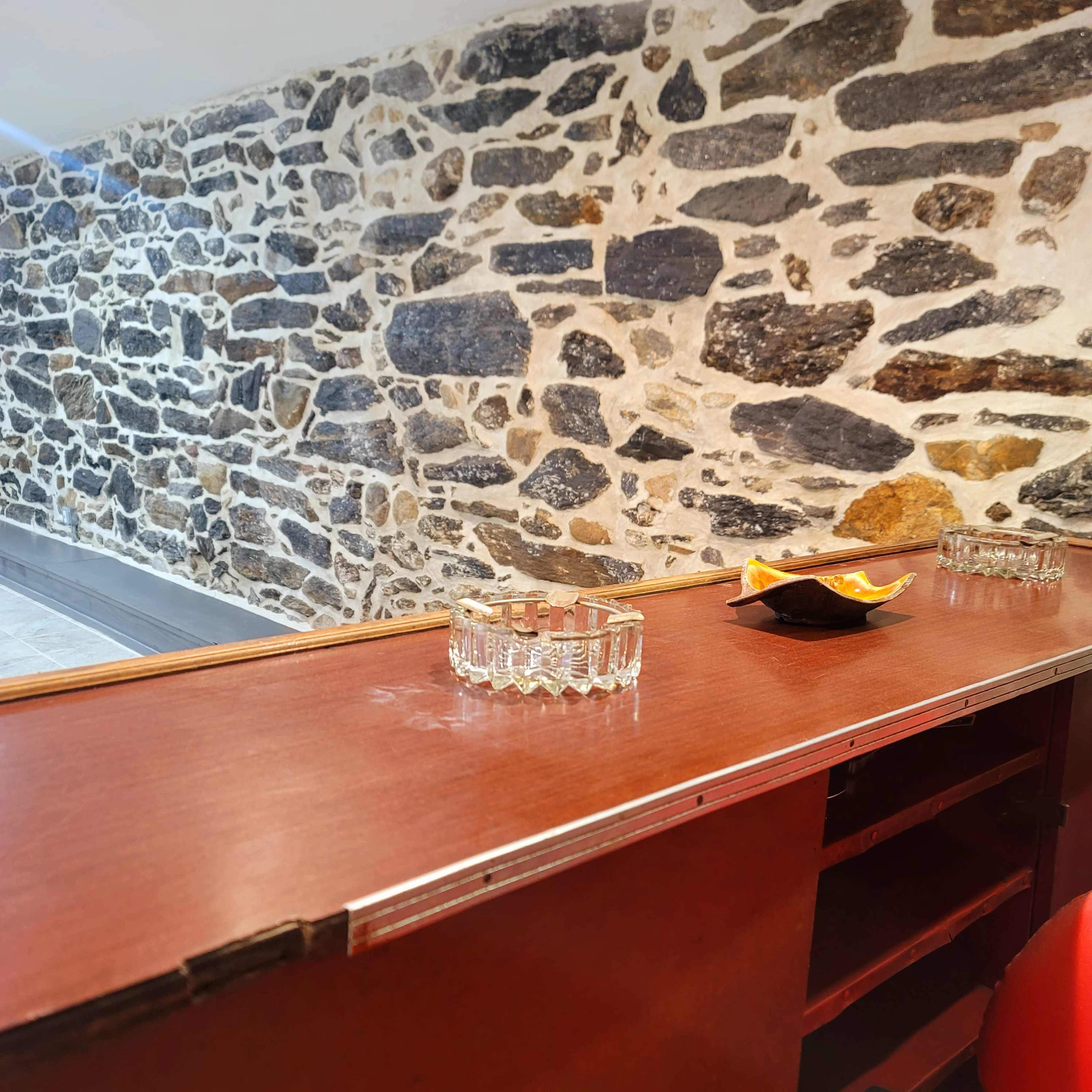 A wooden bar counter with glassware and a dish of snacks overlooks a stone wall in a room.