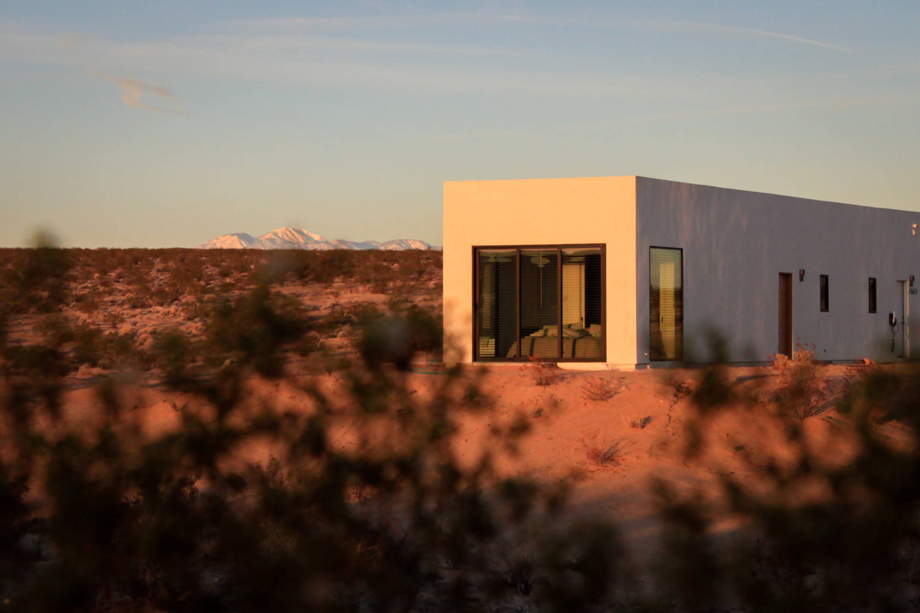 A modern, white rectangular house is set against a backdrop of mountains in a desert landscape at sunset.