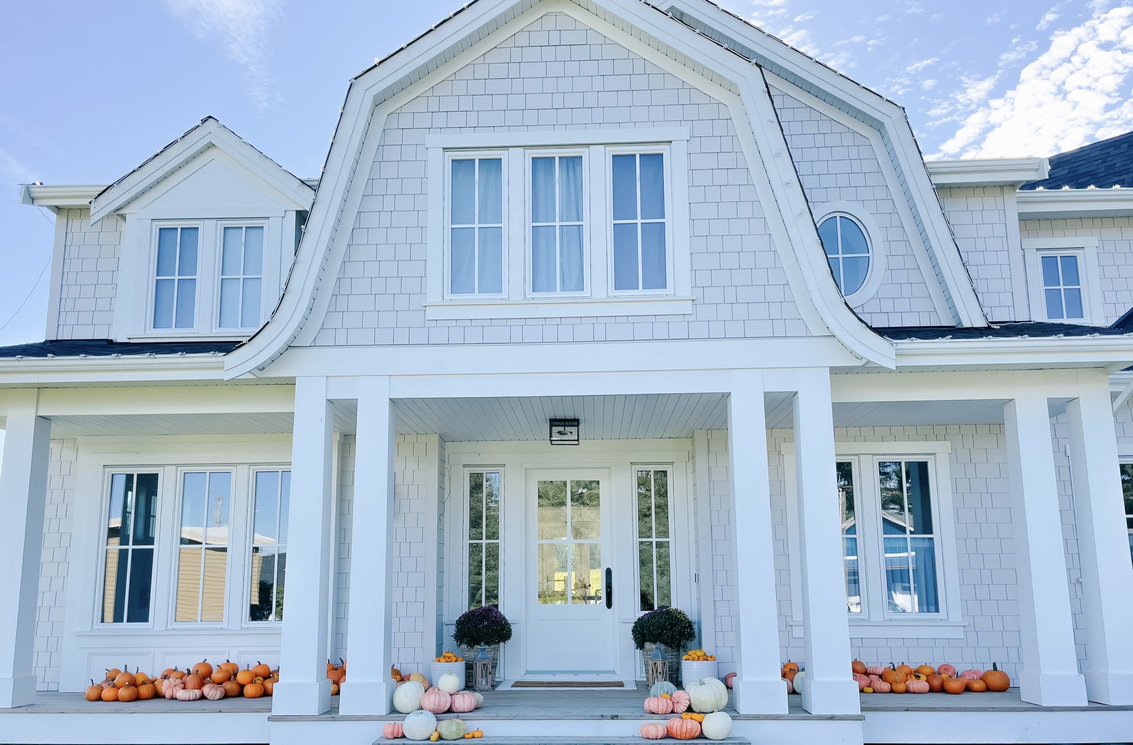 A large, light-colored house features a symmetrical facade with a set of steps leading to a central front door, flanked by white pumpkins and potted plants on the porch.