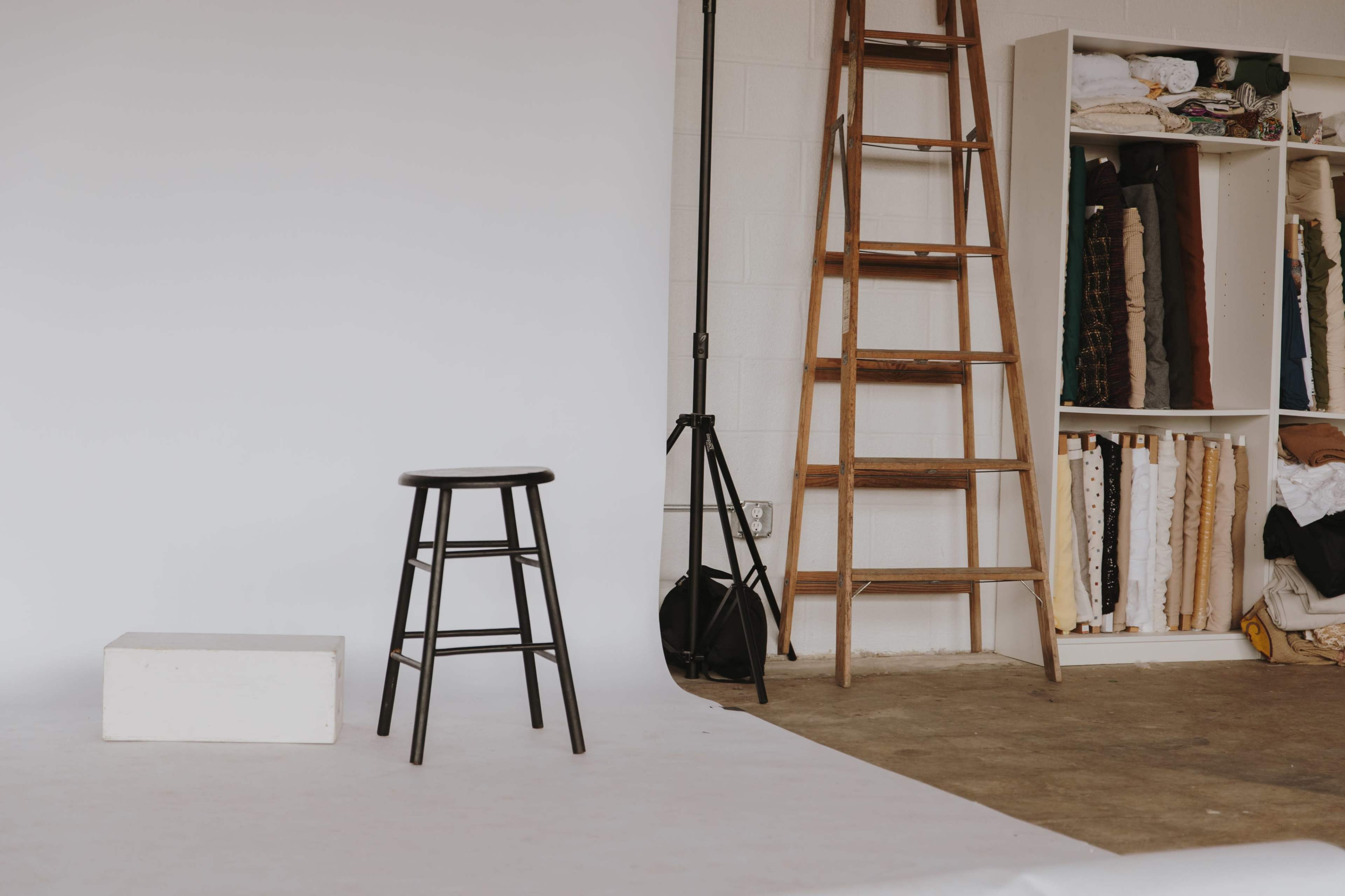 A simple studio setup features a black stool, a white block, a wooden ladder, and shelves filled with fabric.