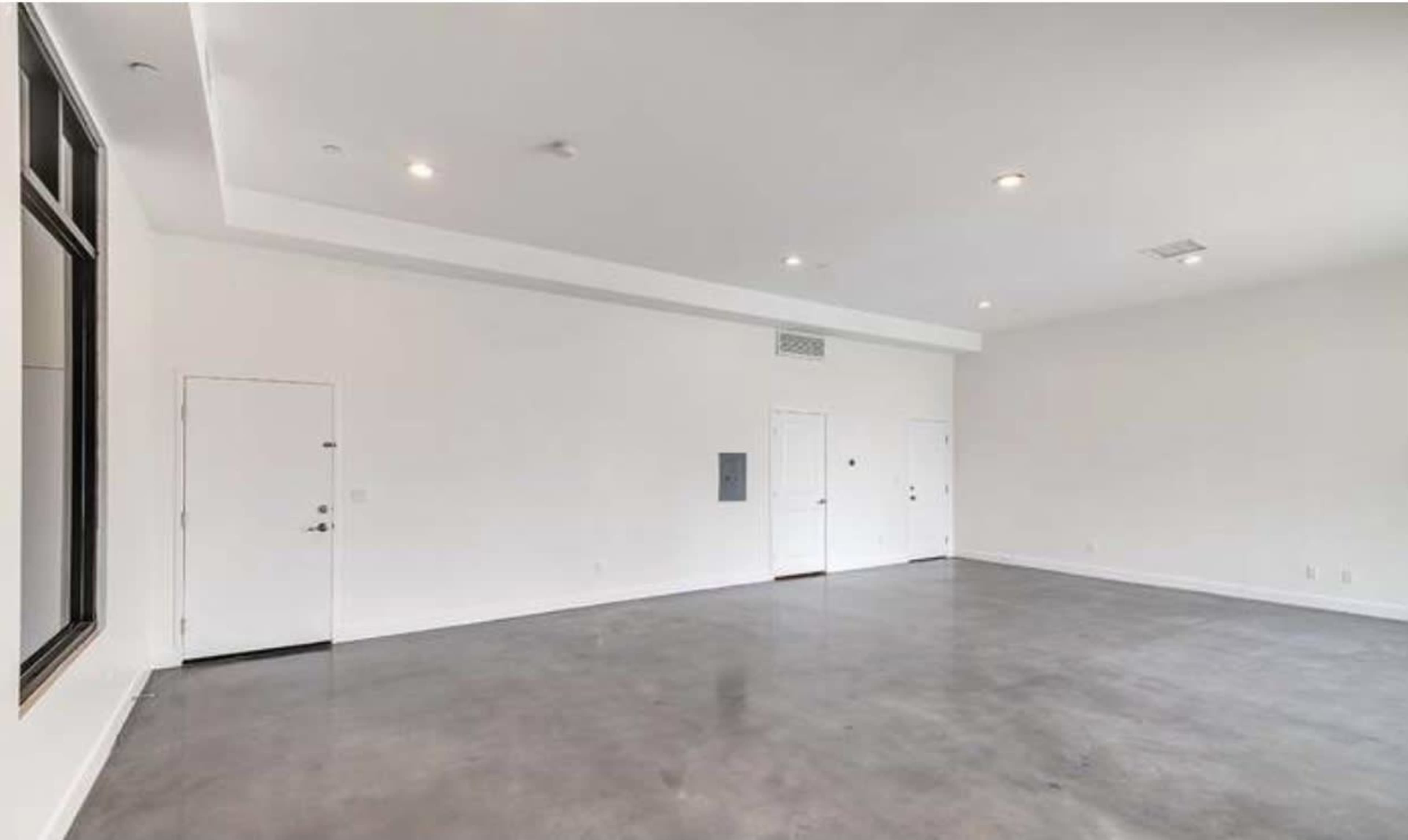 The image shows a spacious, empty room with concrete floors and minimal furnishings, featuring a door and a mail slot on one wall.