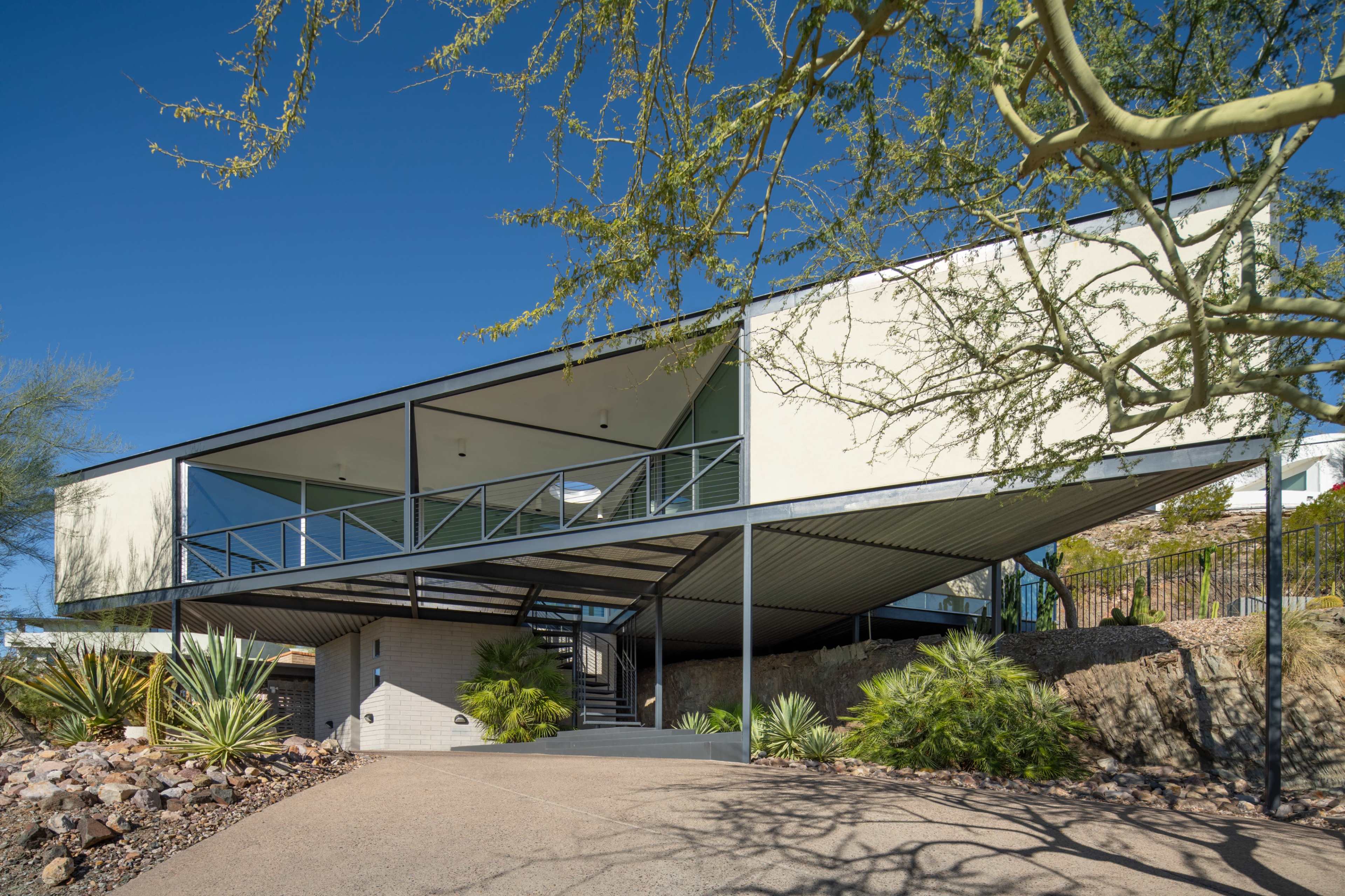 The image shows a modern, elevated house with a distinct angular design, supported by pillars, surrounded by desert landscaping.