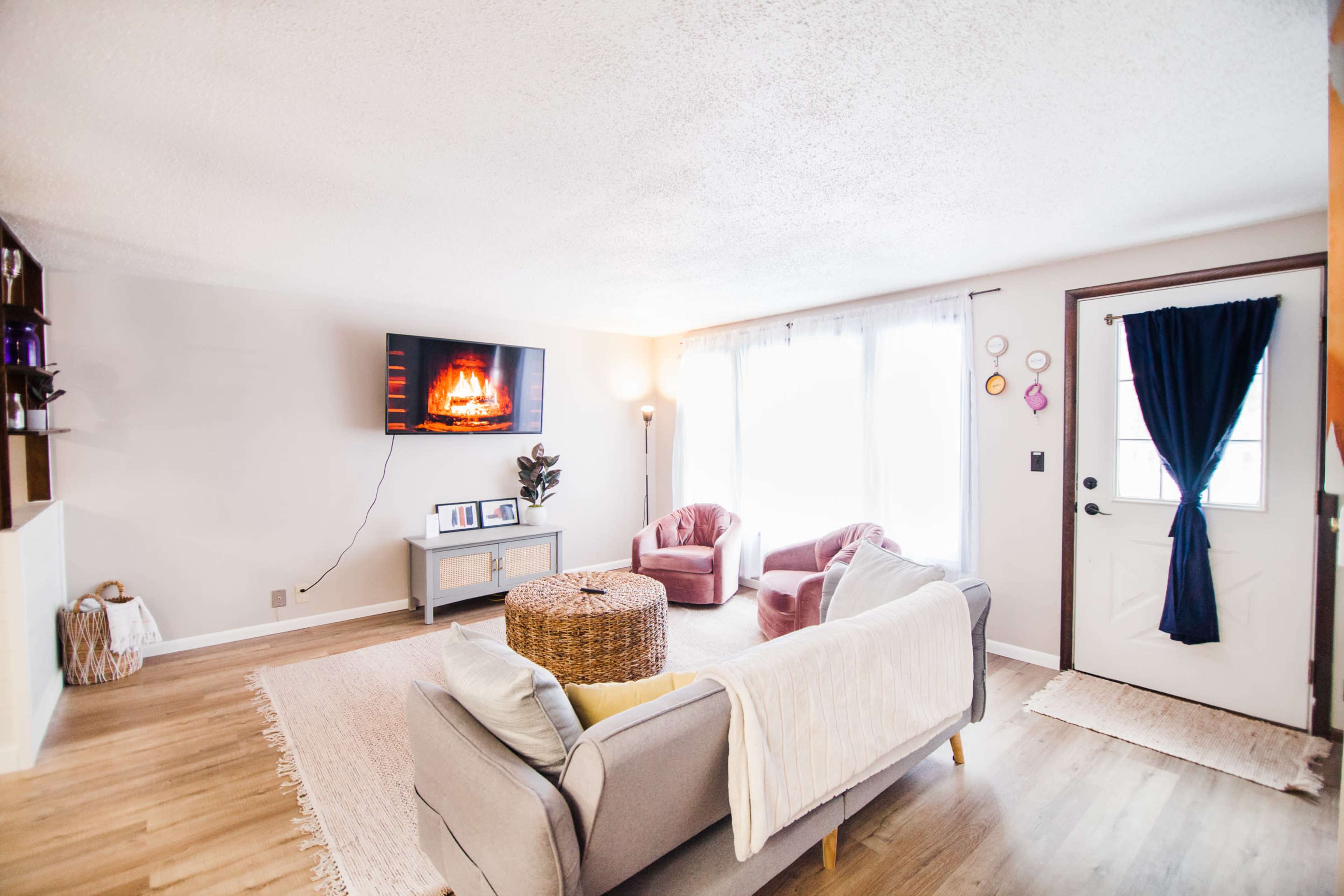 A cozy living room features a sofa, two pink armchairs, a coffee table, and a television displaying a fireplace, with a front door and large windows allowing natural light.
